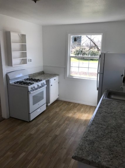 Small kitchen with white walls, featuring a gas stove, oven, refrigerator, and marble-like countertops. A window provides natural light, overlooking a grassy area. Wooden floors and open shelving are visible.