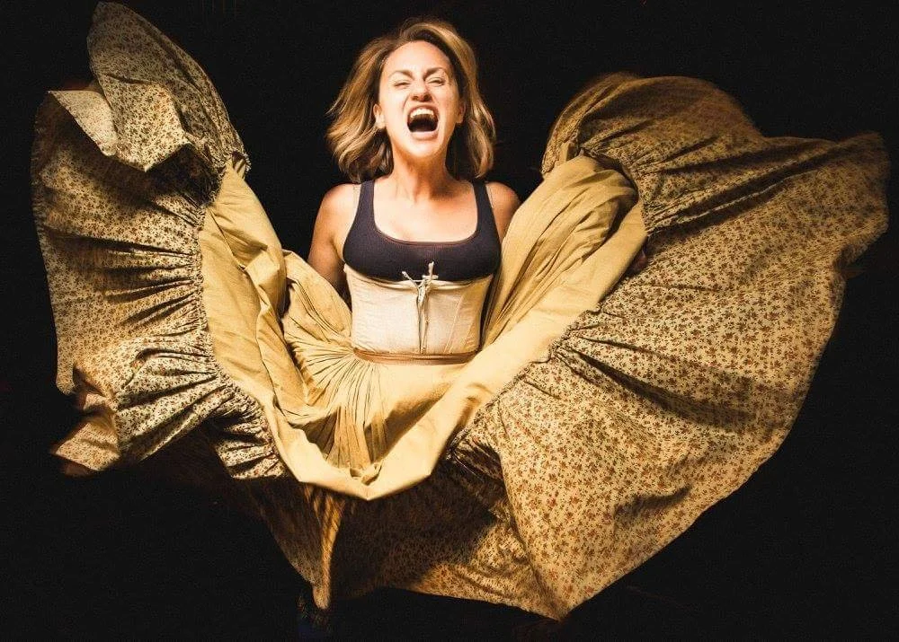Woman with blonde hair wearing a corset and a flowing skirt, energetically posing with an open mouth in an expression of excitement, against a dark background.