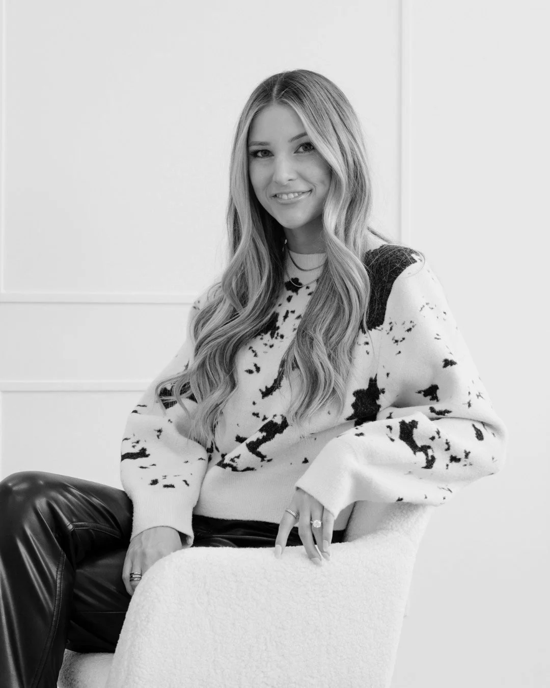Black and white photo of a woman with long hair, wearing a patterned sweater, sitting on a chair with crossed legs.