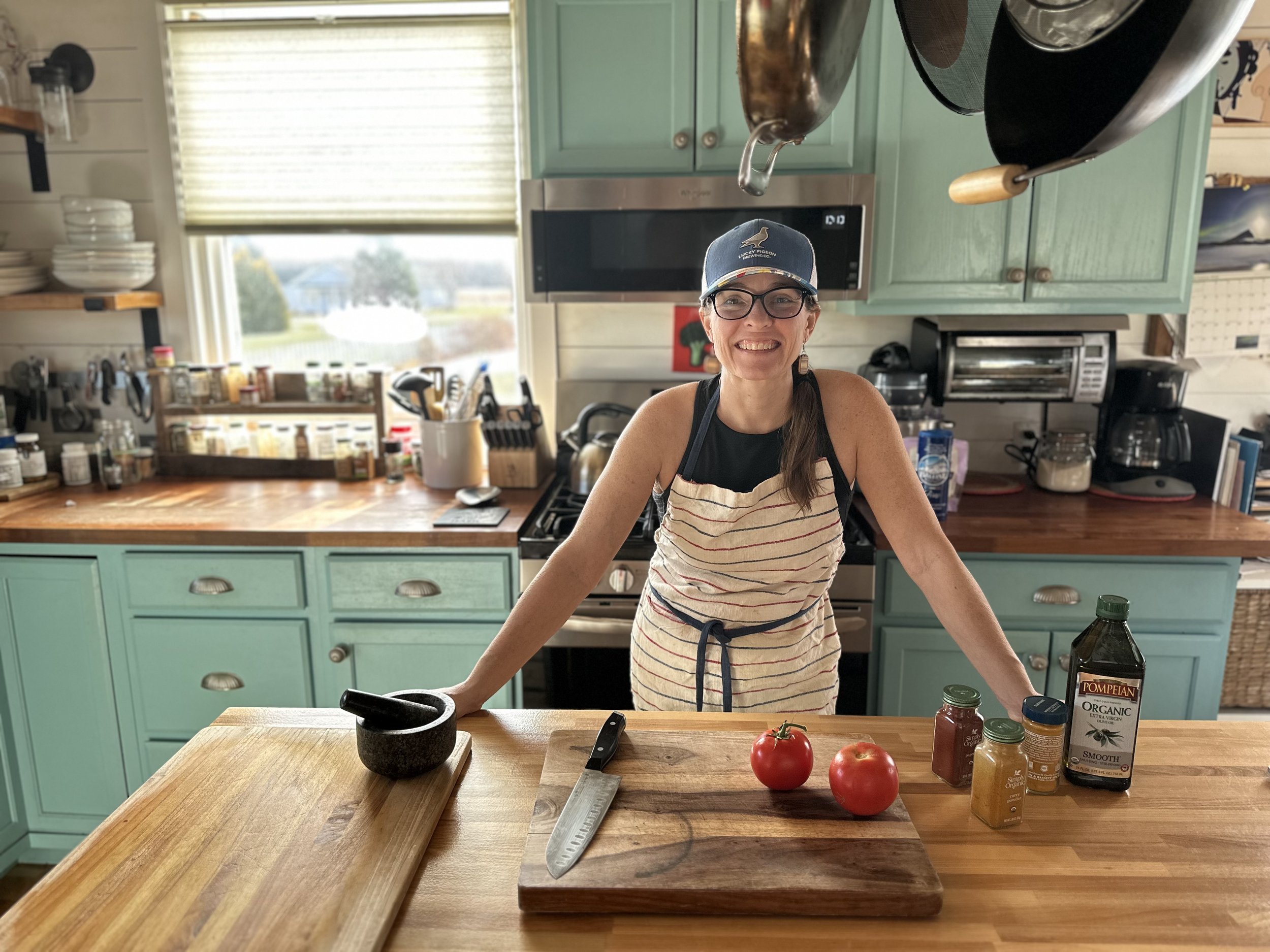 An image of a woman smiling with a hat and an apron on in front of a counter with a knife and some tomatoes