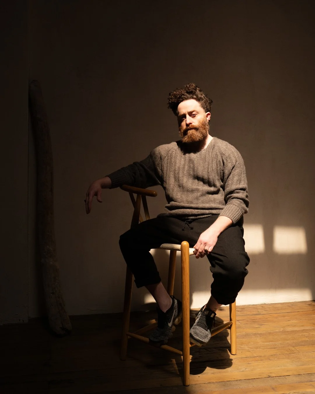 A man with a full beard and curly hair sitting on a wooden chair in a dimly lit room. He is wearing a gray sweater, black pants, and sneakers. Shadows and light create a moody atmosphere. Sruli Recht
