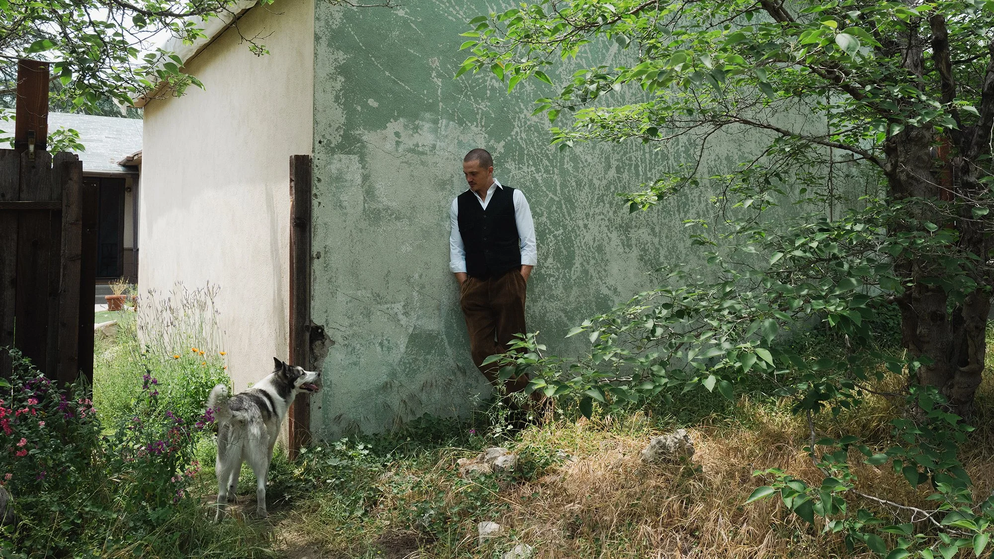 Karl Glusman leaning against a weathered green wall in a garden, with a Siberian Husky dog nearby, surrounded by trees and plants.