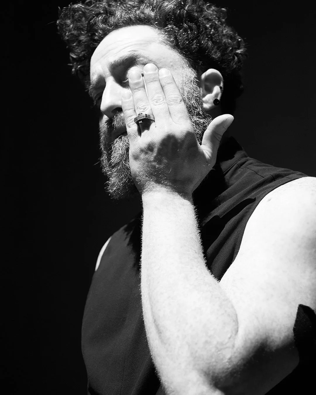 A black and white photo of a man with a beard and curly hair covering his face with his hand, wearing rings and earrings. Sruli Recht