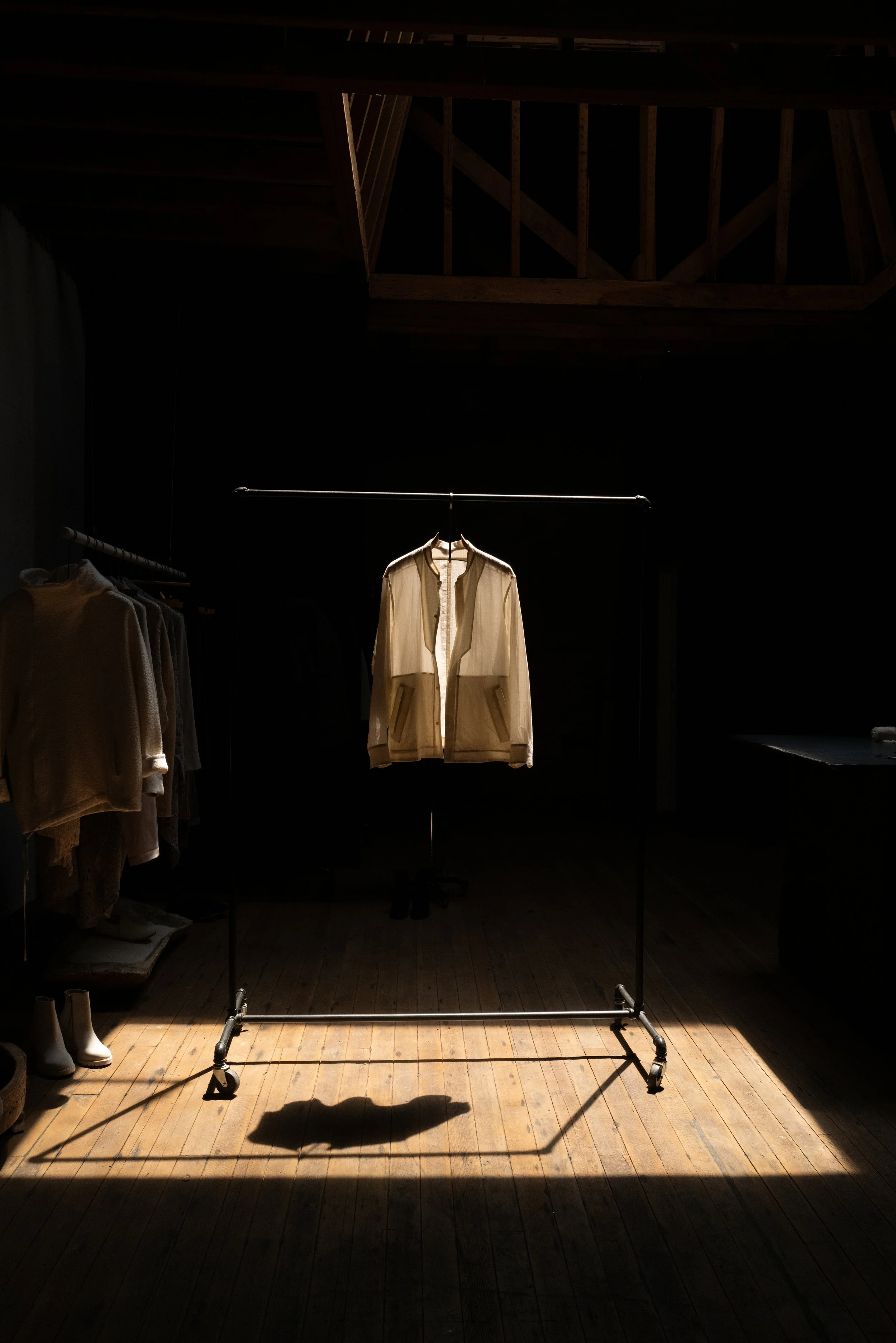 A clothing display with a single cream-colored shirt hanging on a metal rack, illuminated by a focused light in a dark room with wooden floors. Eyn Vas
