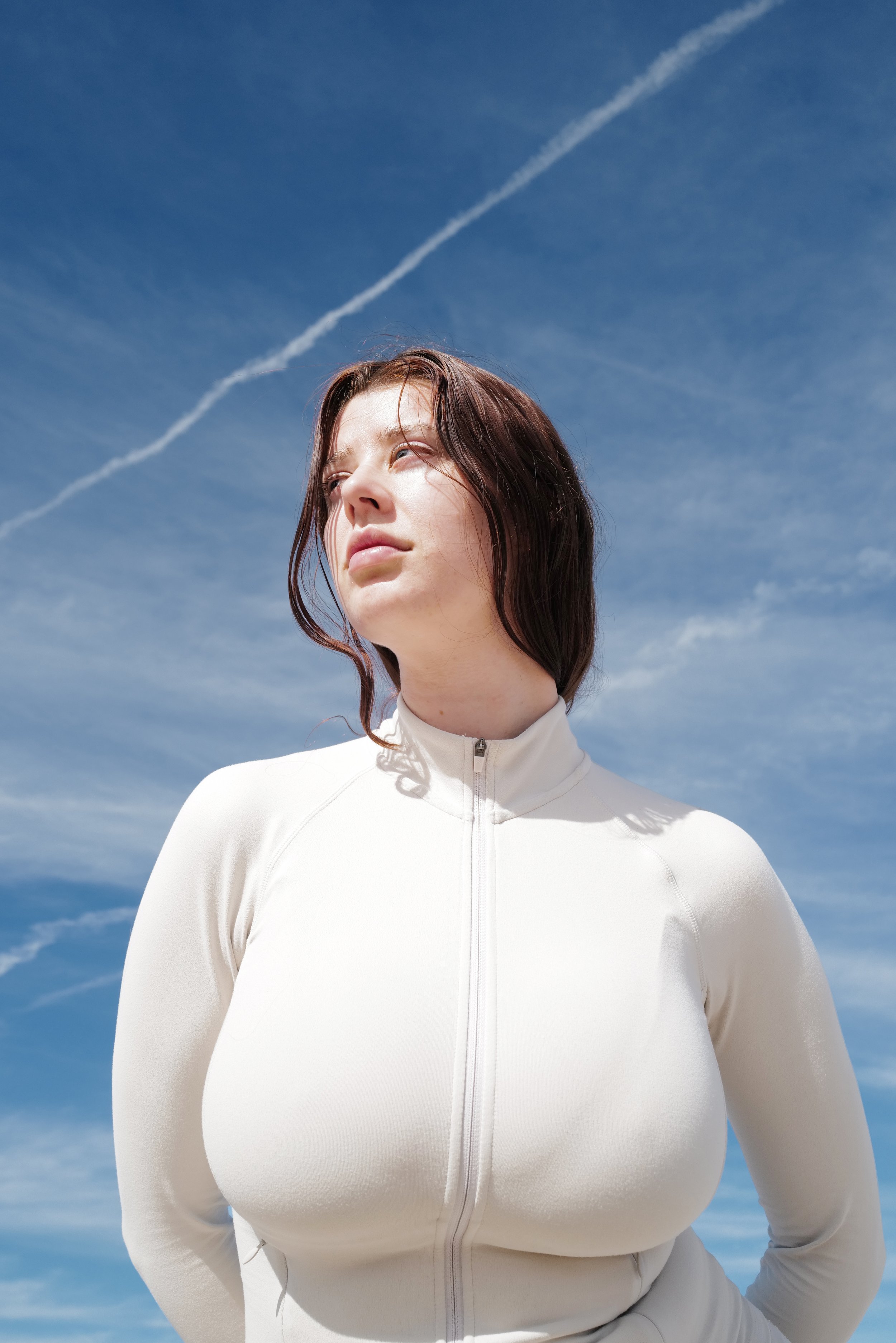 Sarah McDaniel is outside against a blue sky with wispy clouds and a contrail. She is wearing a white zip-up jacket and looking slightly upward.