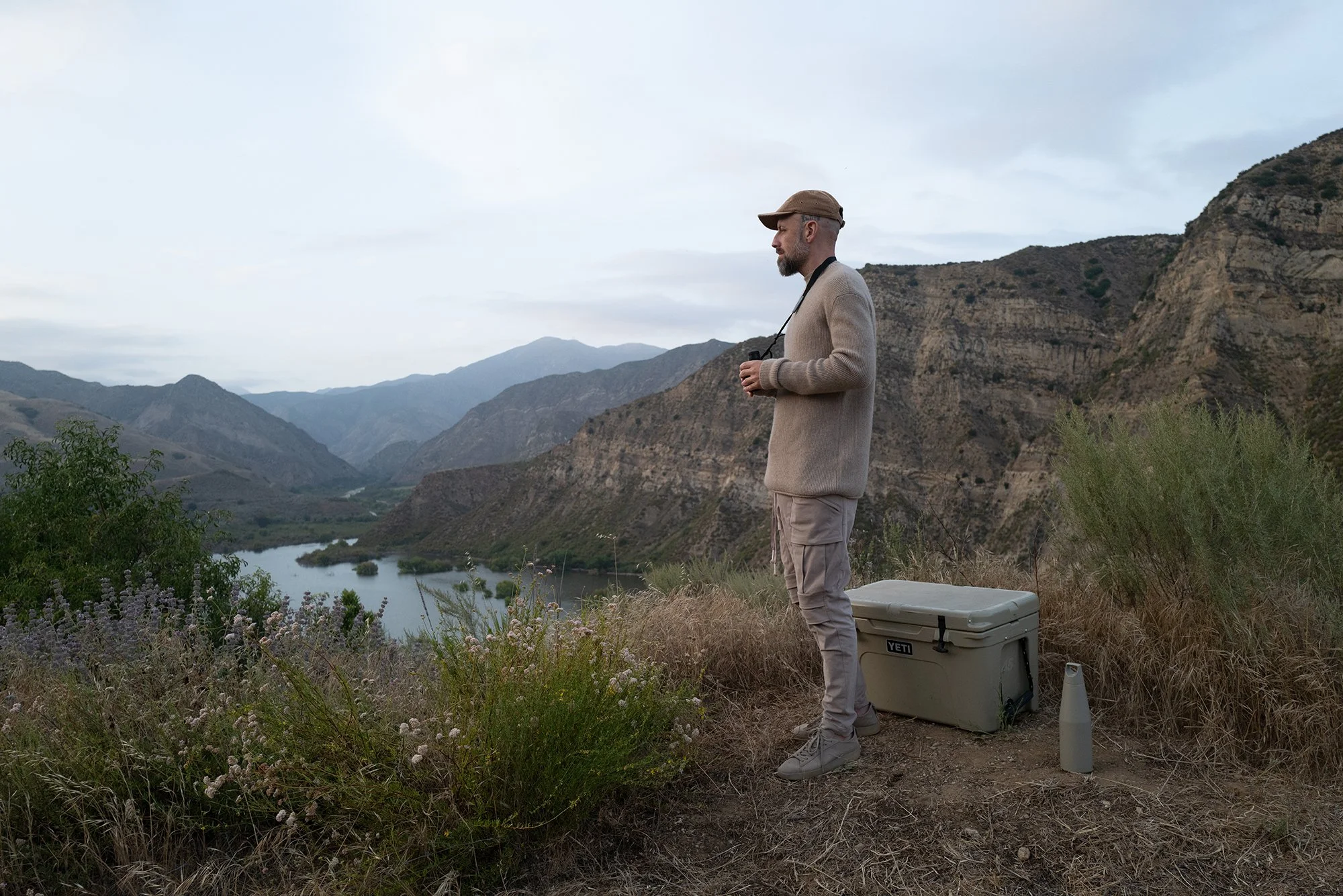 A man standing on a hillside overlooking a river and rugged mountains during overcast weather, holding a camera and wearing outdoor clothing. Jona Sees. Eyn Vas