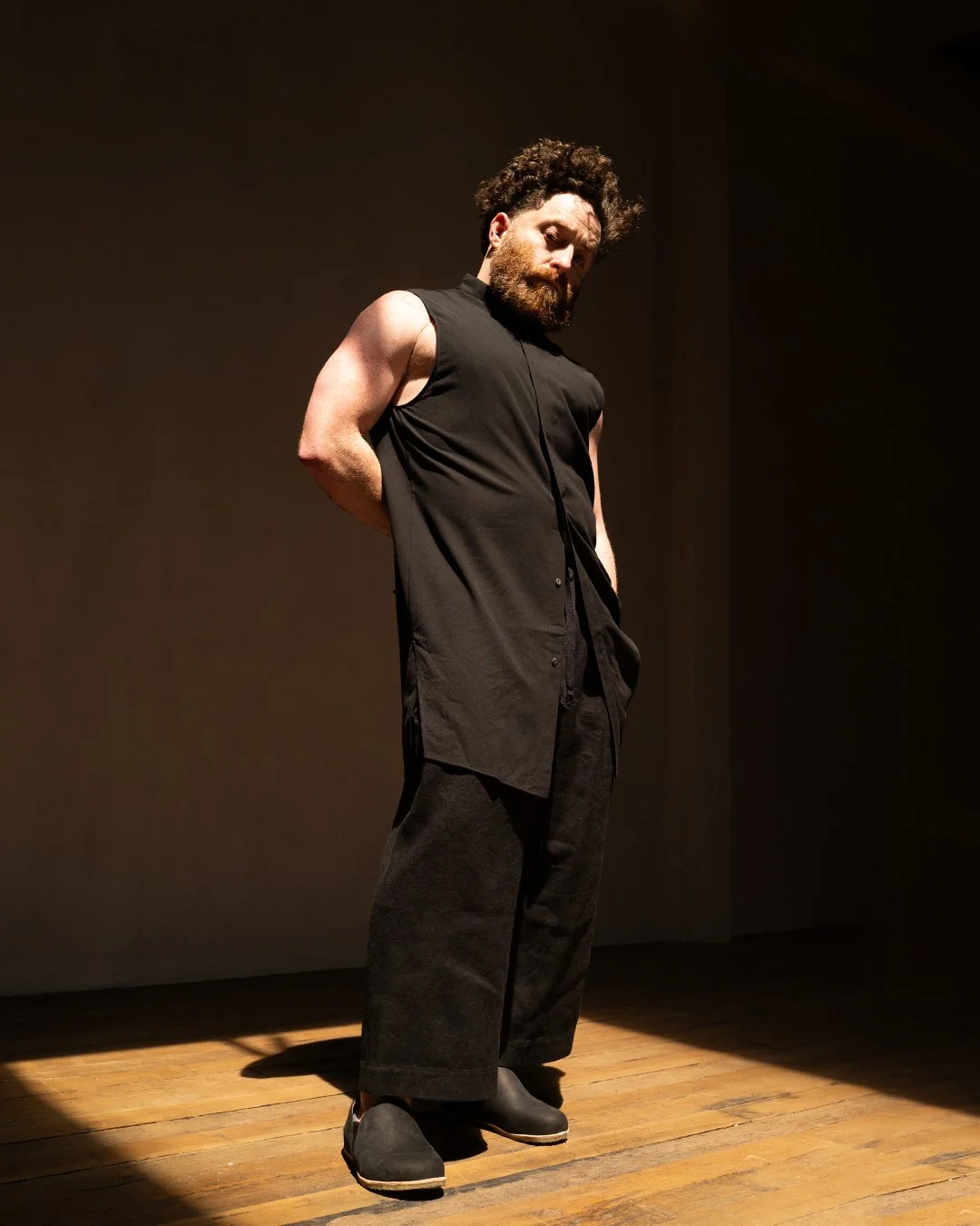 A man with dark, curly hair and a beard stands on a wooden floor against a beige wall, wearing a sleeveless black shirt, loose black pants, and black shoes, with his hands behind his back, looking down. Sruli Recht