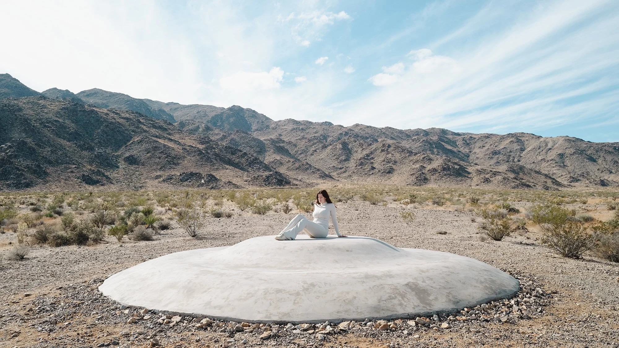 A woman dressed in white sitting on a white rock formation in a desert landscape with mountains in the background and a partly cloudy sky. Sarah McDaniel