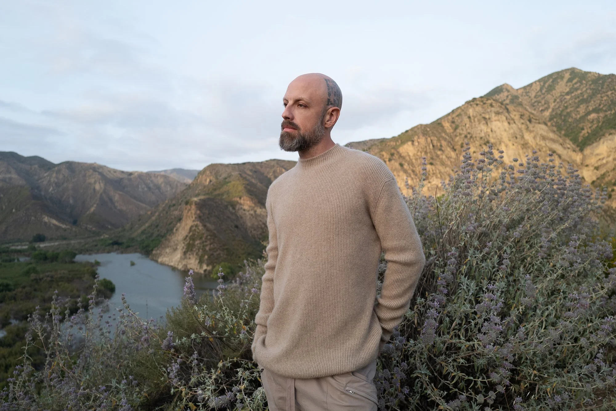 Jona Sees, the fashion designer is standing outdoors near purple wildflowers, overlooking a river with mountains in the background during daylight. Jona Sees. Eyn Vas