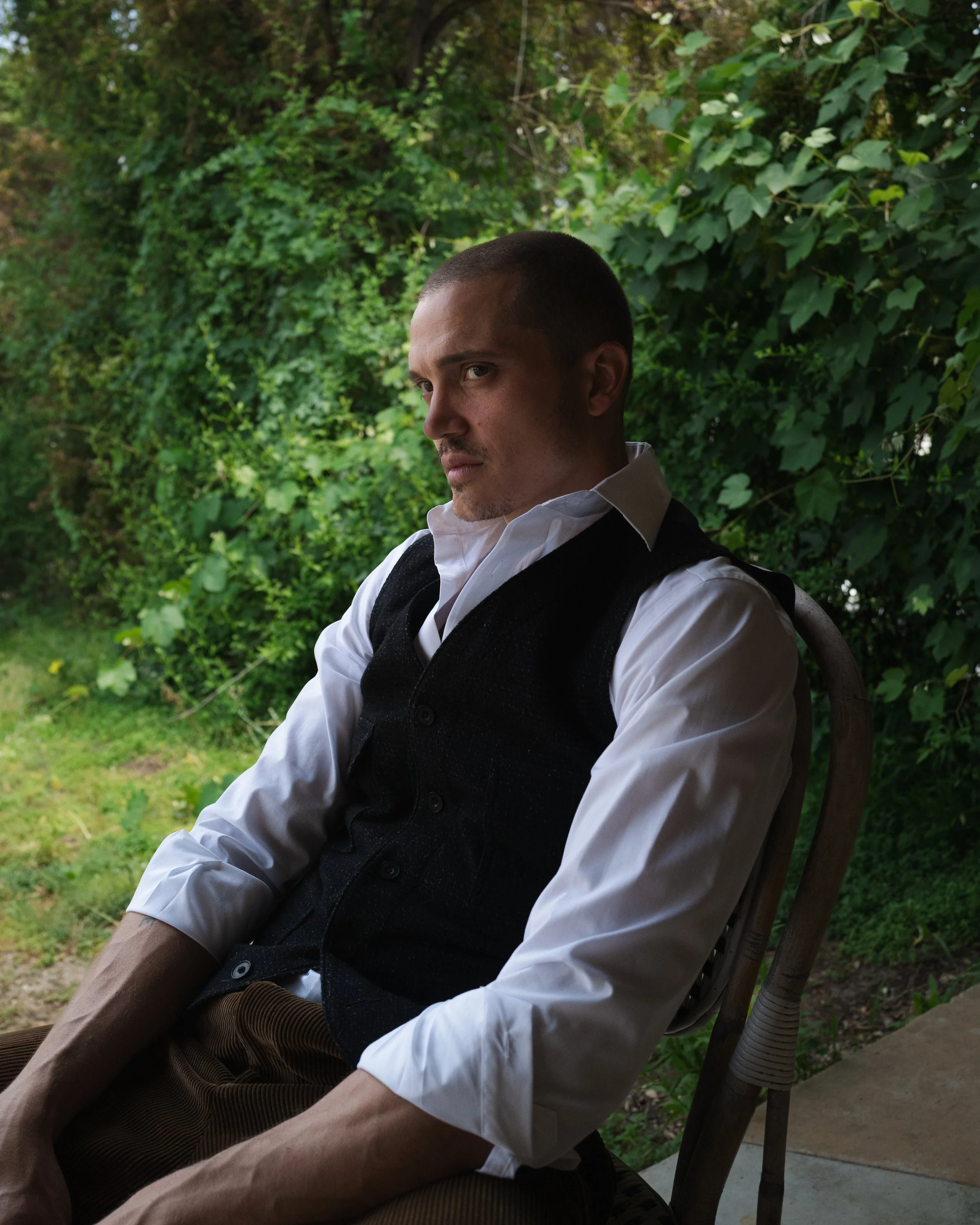 A man with a shaven head and serious expression, dressed in a white shirt and dark vest, sitting outdoors with greenery in the background. Karl Glusman