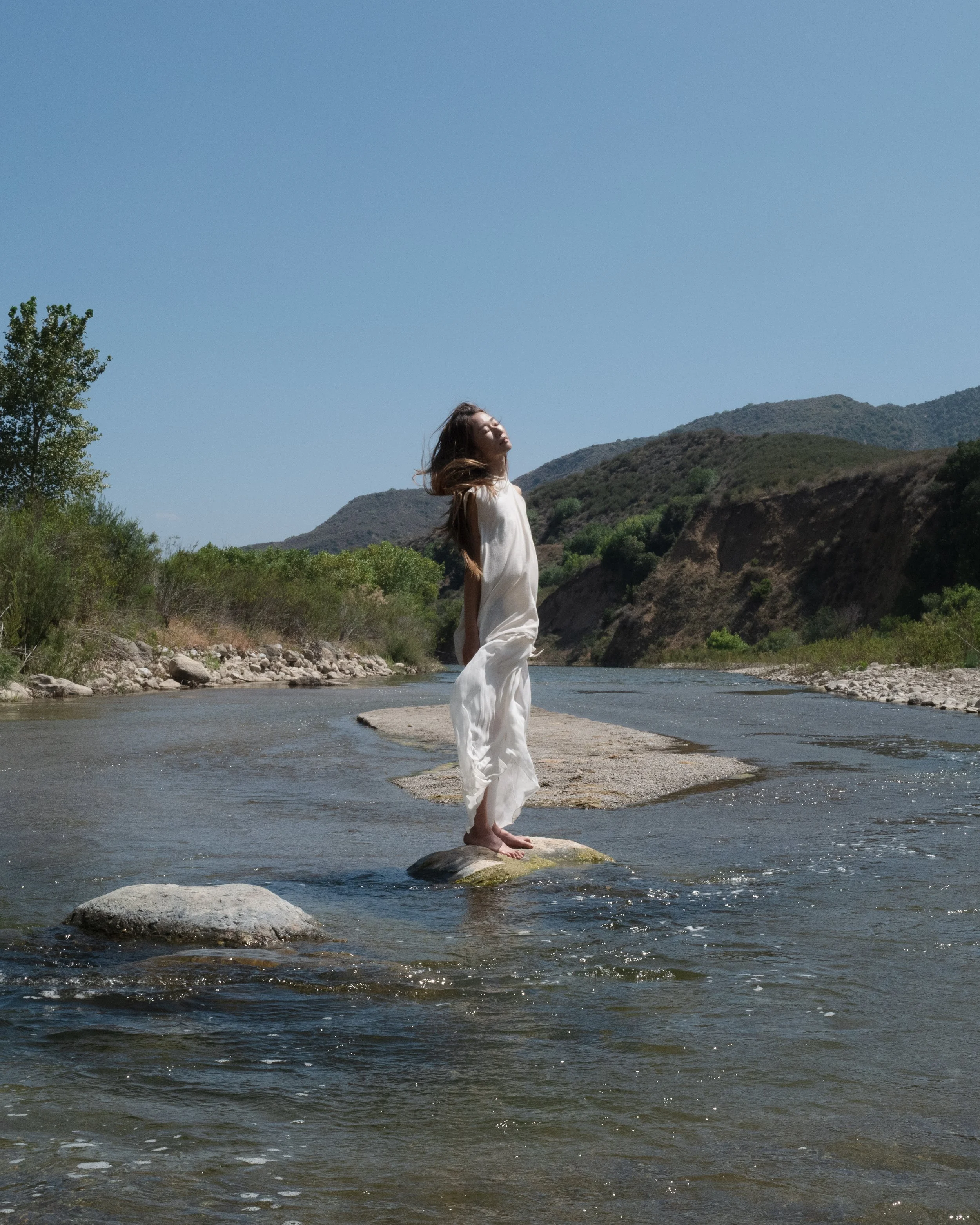 A woman stands on a rock in a river with her eyes closed, wearing a white dress, with mountains and greenery in the background under a clear blue sky. Eyn Vas