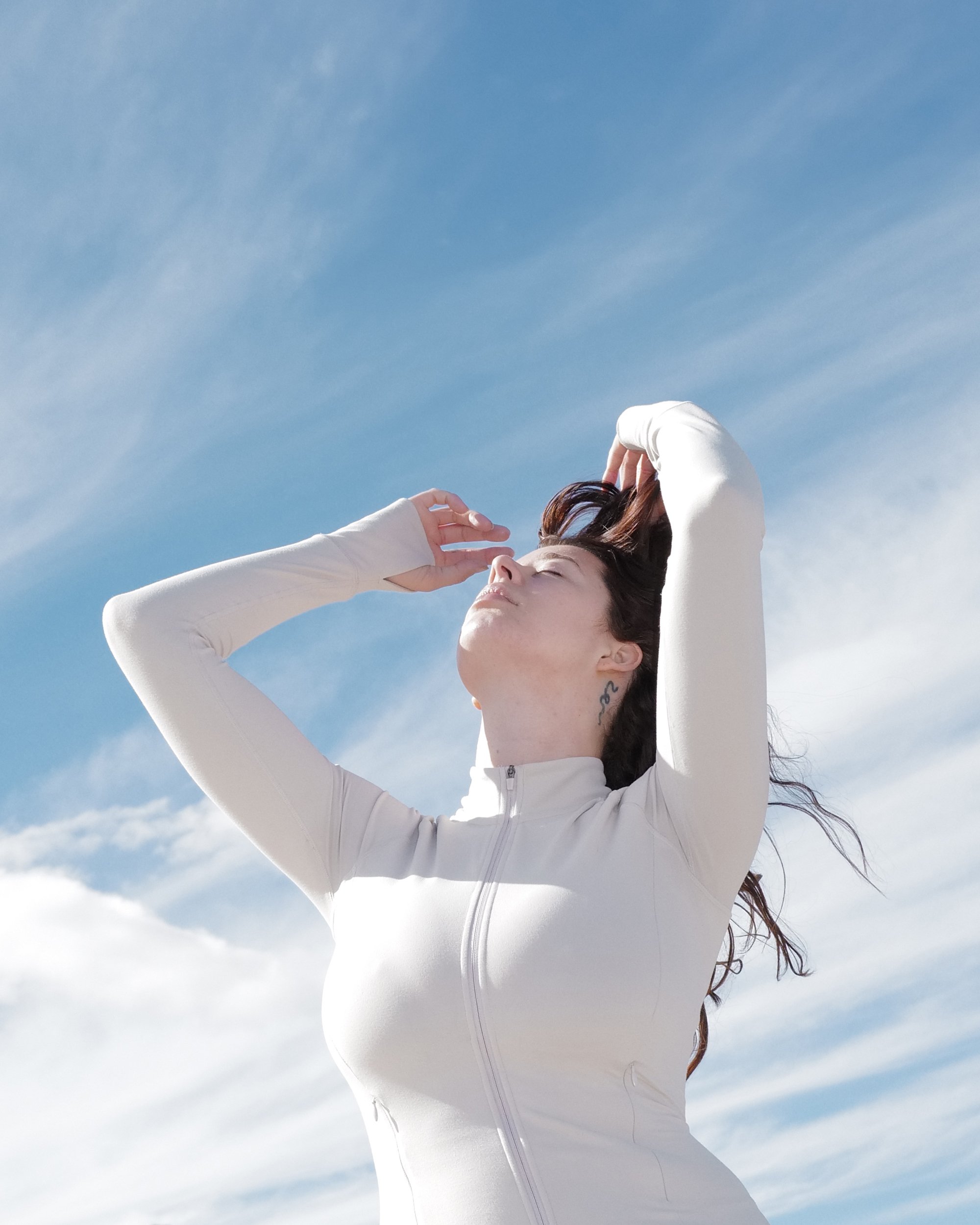 A woman with dark hair and a small tattoo on her neck, wearing a white zip-up top, standing outdoors under a blue sky with clouds, with her eyes closed, raising her left hand to her forehead. Sarah McDaniel