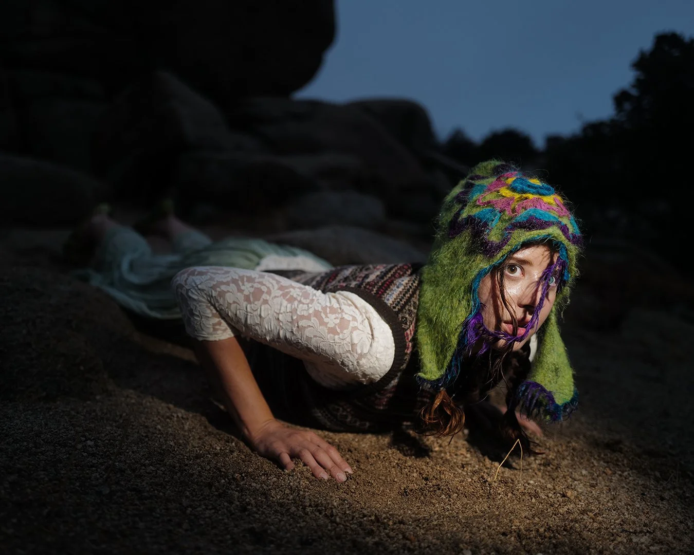 Krotchy wearing a colorful knitted hat, lies on the ground on her stomach, supporting herself with her hands, in a dark outdoor setting.