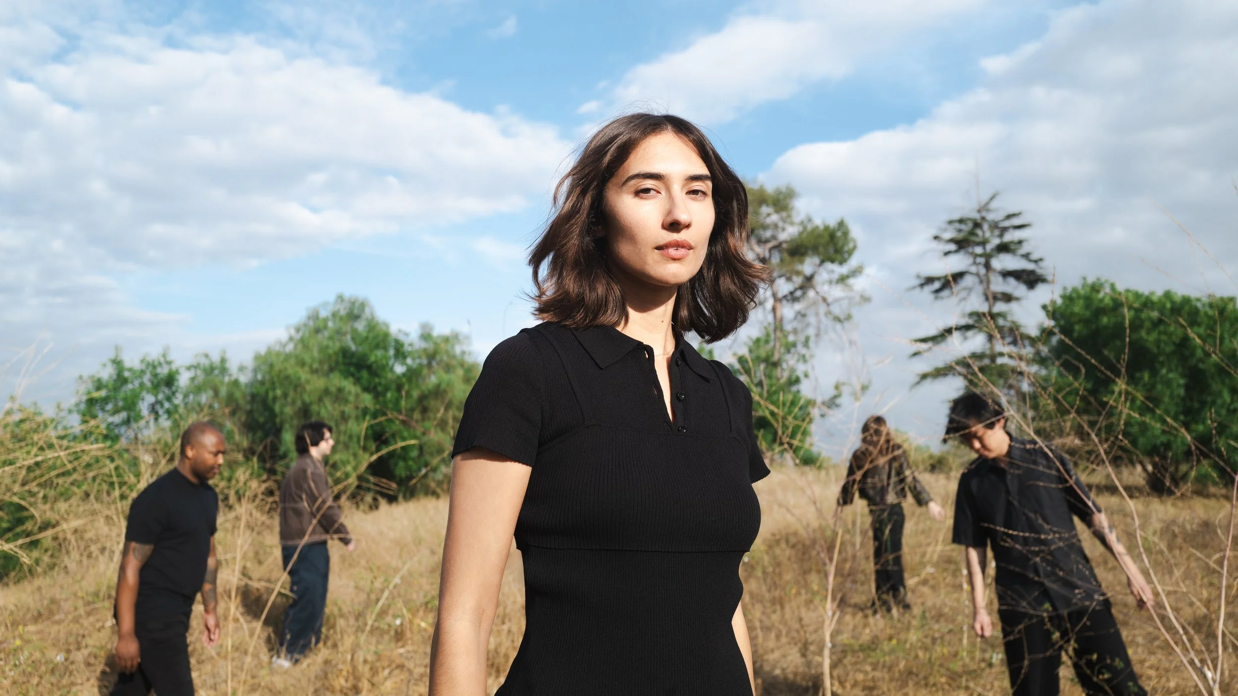 A woman with shoulder-length brown hair wearing a black shirt in an outdoor setting with four other people in the background, walking through nature with trees and blue sky with clouds.
