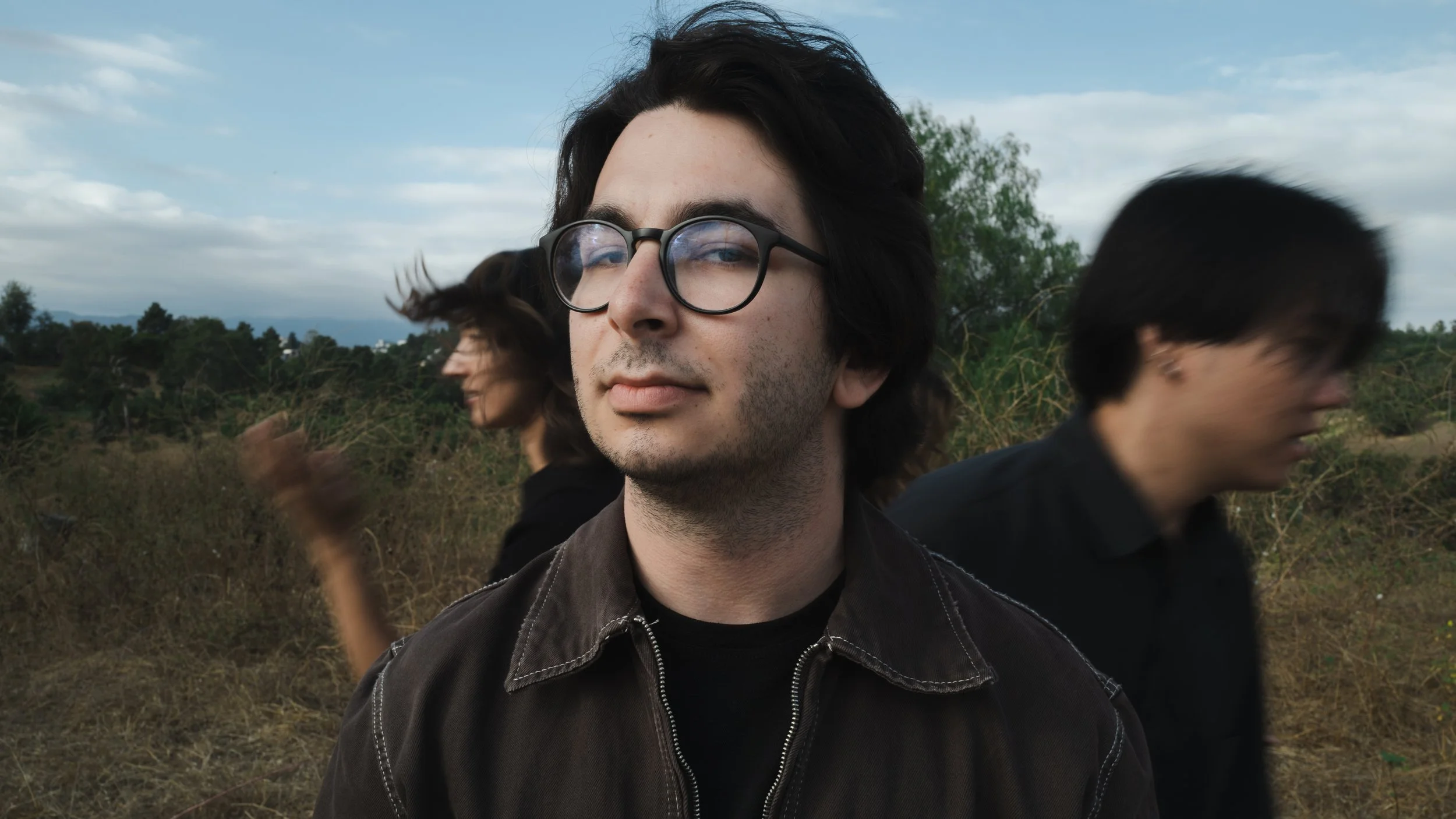 A young man with dark hair, glasses, and a slight stubble stands outdoors with two women blurred behind him, in a field with trees and a cloudy sky.