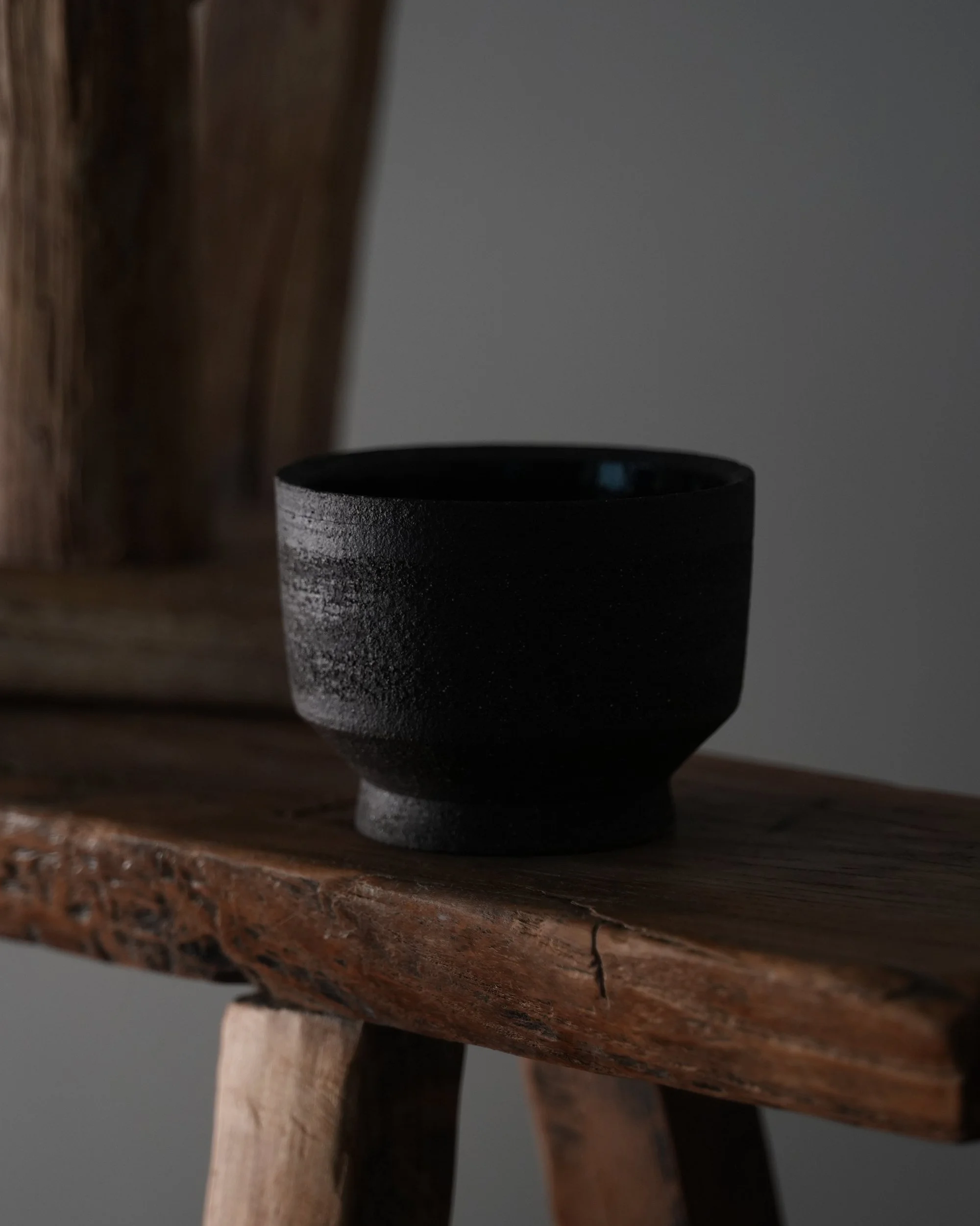 Close-up of a black ceramic cup on a rustic wooden surface with a dark background. Akanendo