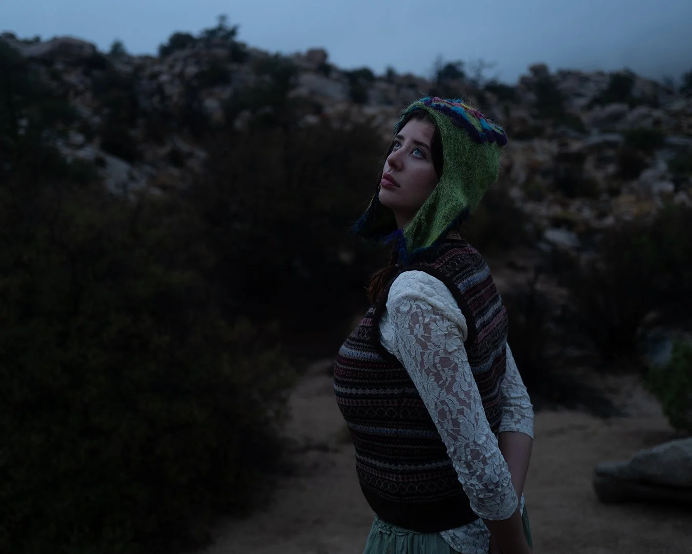 A young woman standing outdoors at dusk, wearing a colorful knit hat with ear flaps and a patterned vest over lace sleeves, looking upwards with a thoughtful expression. Sarah McDaniel. Krotchy