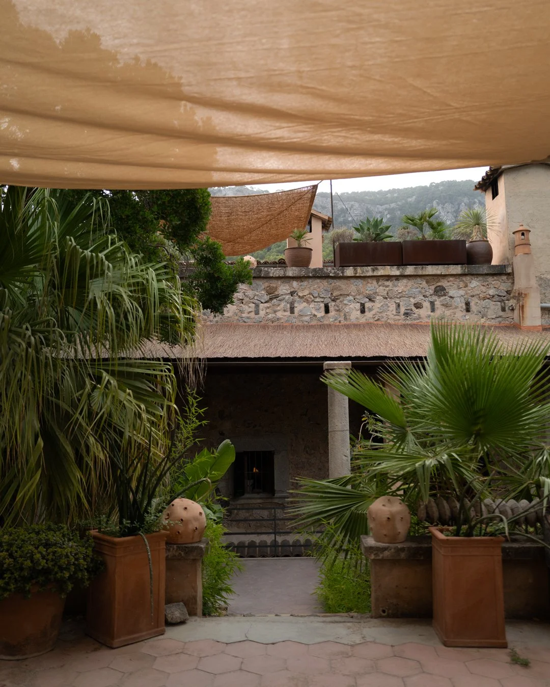 A courtyard with potted tropical plants and cacti, surrounded by stone and stucco walls, with hills in the background. Canvas shades hang overhead. Hotel Corazon. Deia, Mallorca