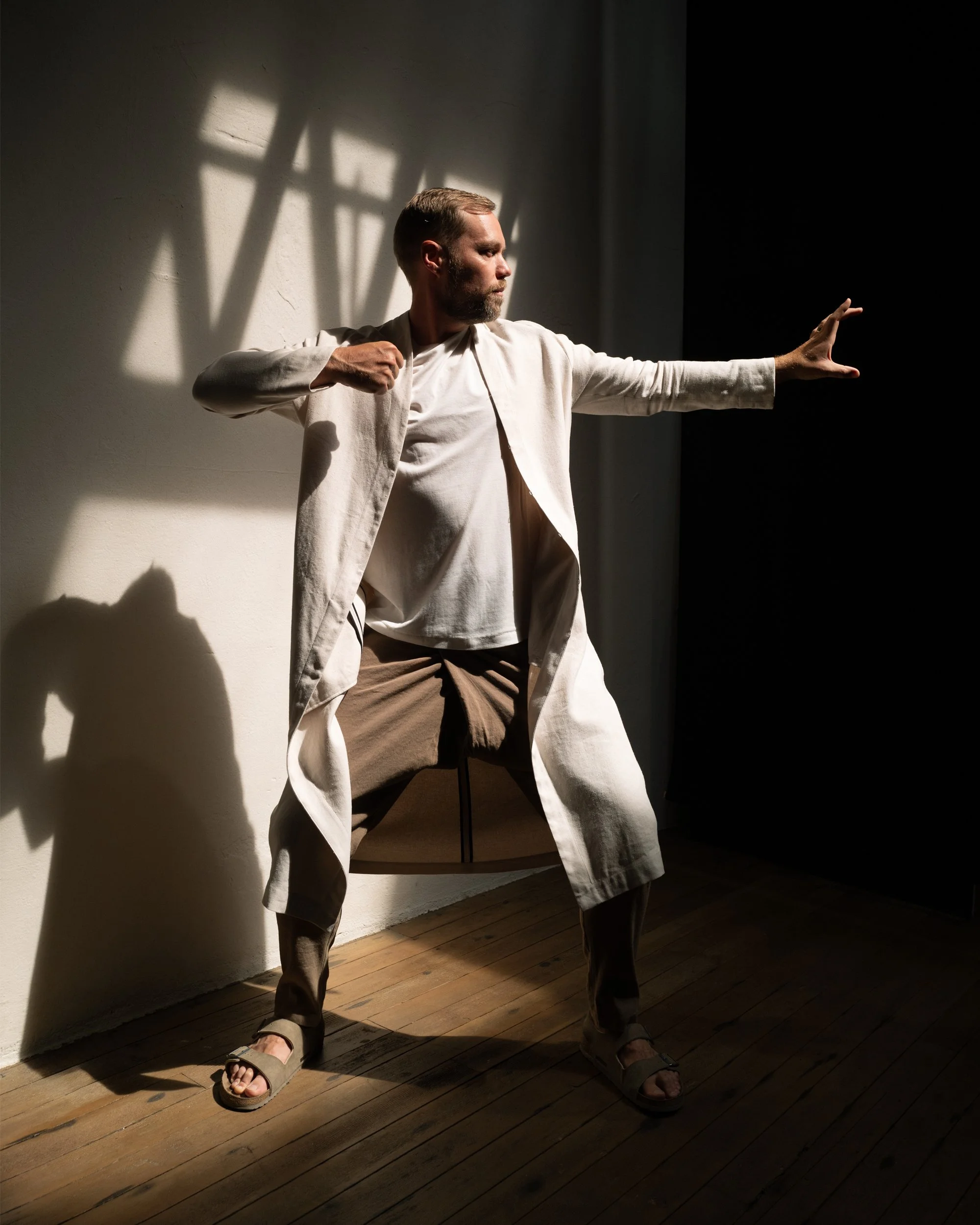 A man with a beard and short hair poses against a wall with light and shadow patterns, wearing loose beige clothing and sandals, reaching out with one arm. Eyn Vas