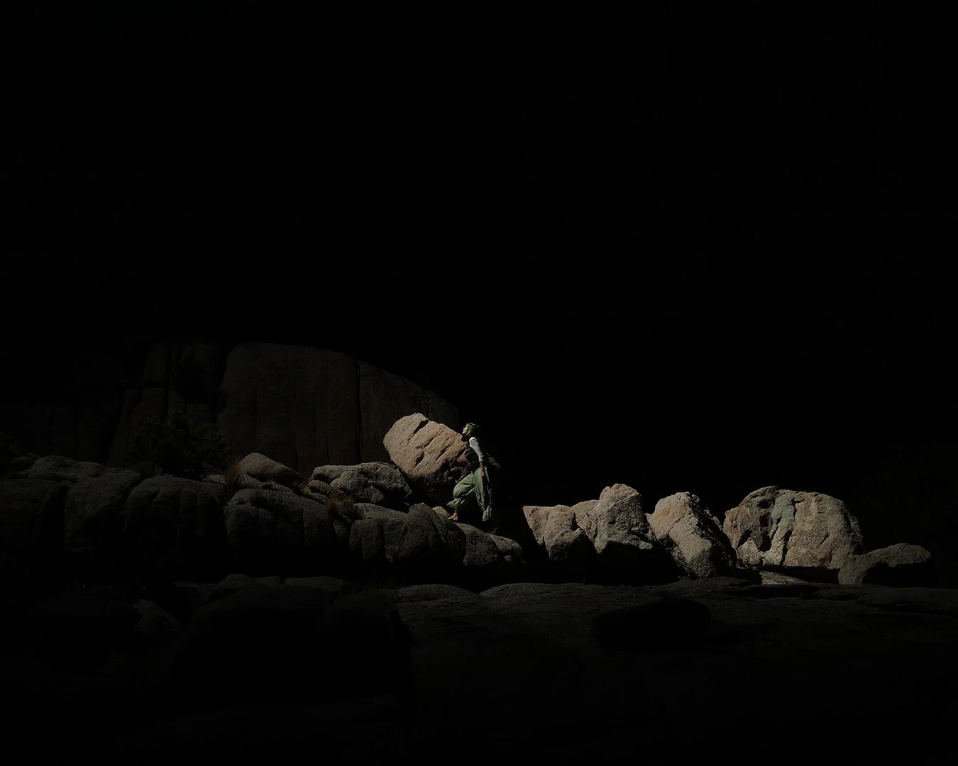 A person with a backpack climbing over rocks in a dark environment, with minimal lighting highlighting the rocks and the climber. Sarah McDaniel. Krotchy