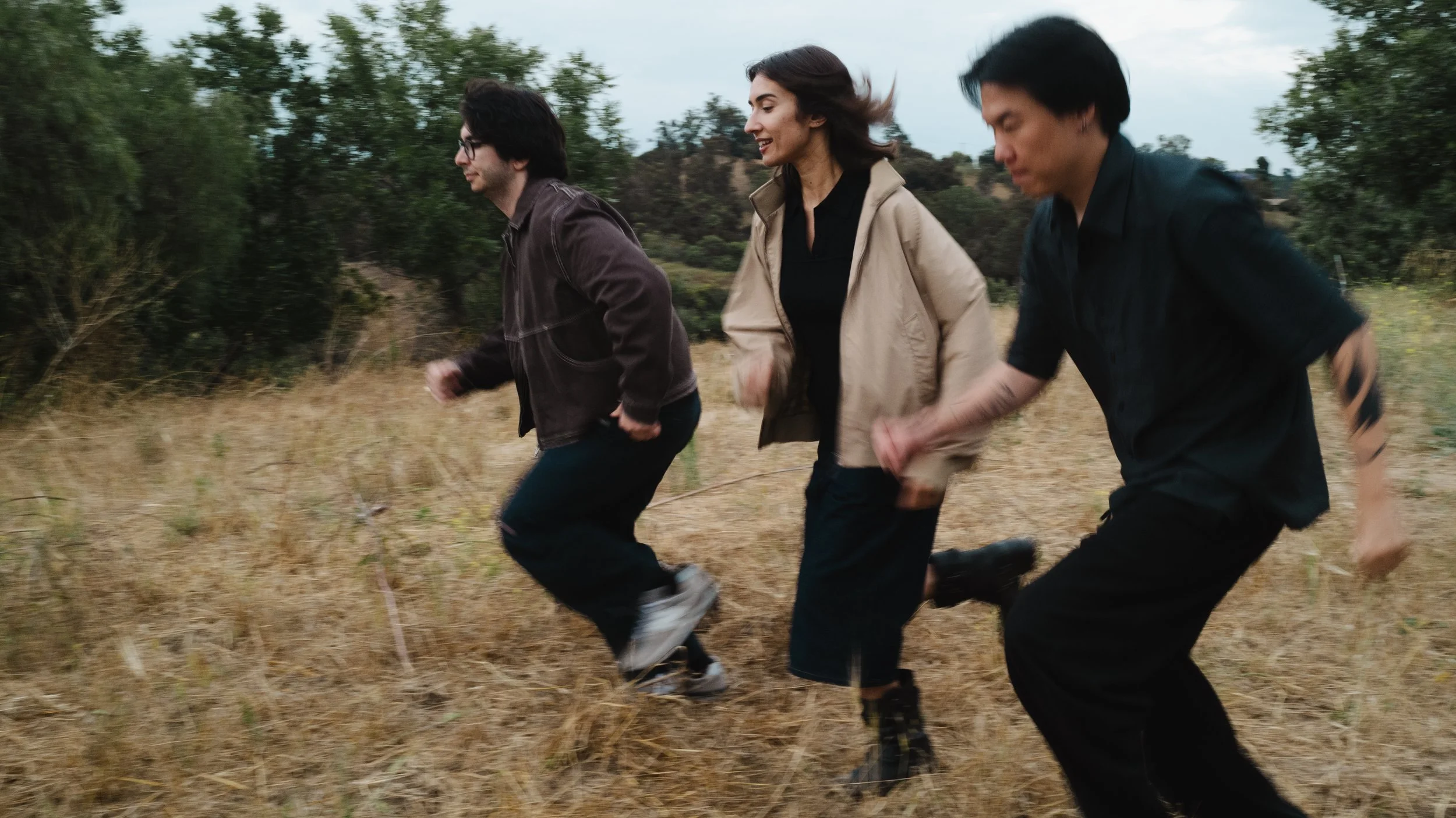Three people running outdoors in a grassy field with trees in the background.