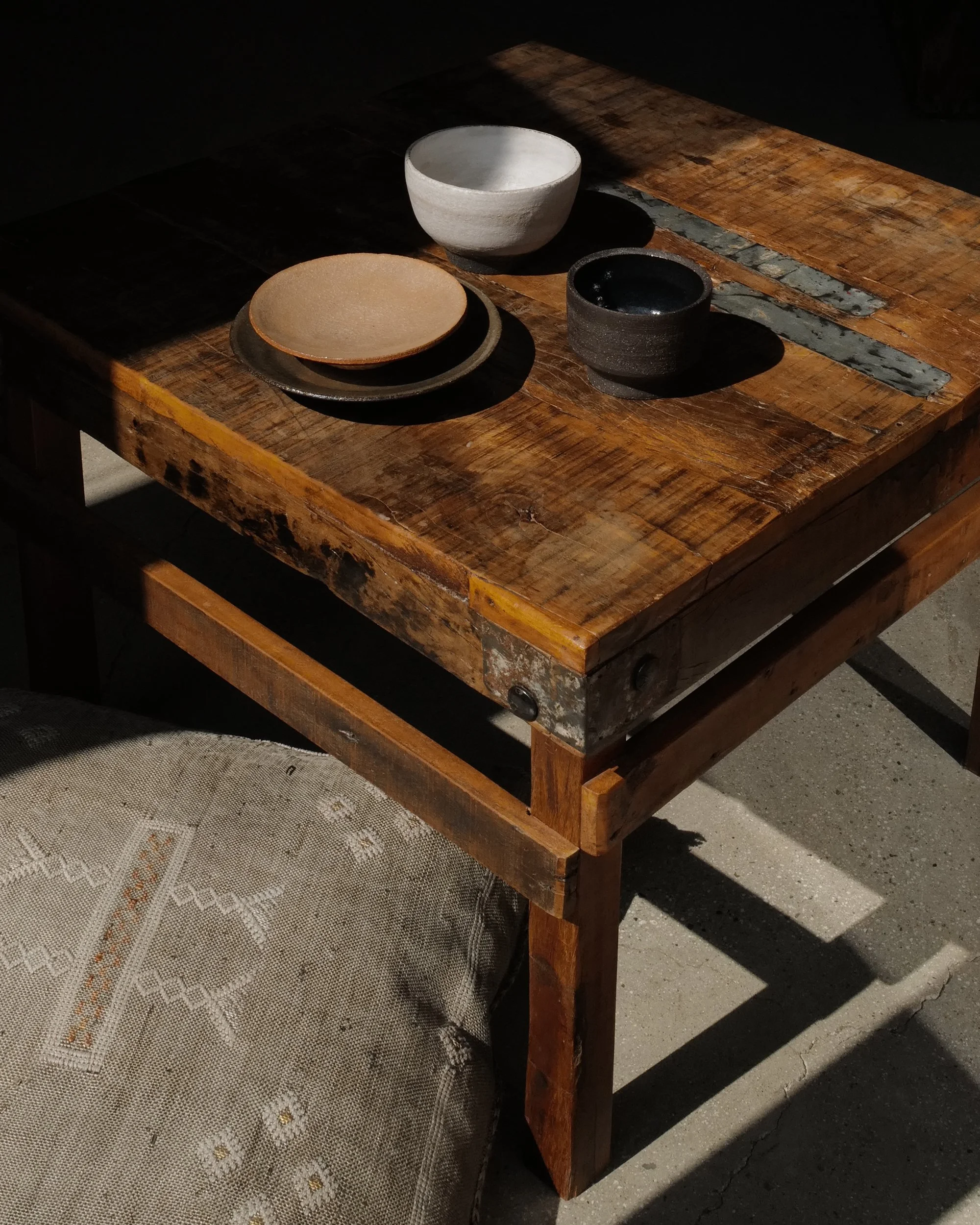 A rustic wooden table with four ceramic bowls and plates on it, with a lace tablecloth partially visible underneath. The table is set outdoors with sunlight casting shadows. Akanendo