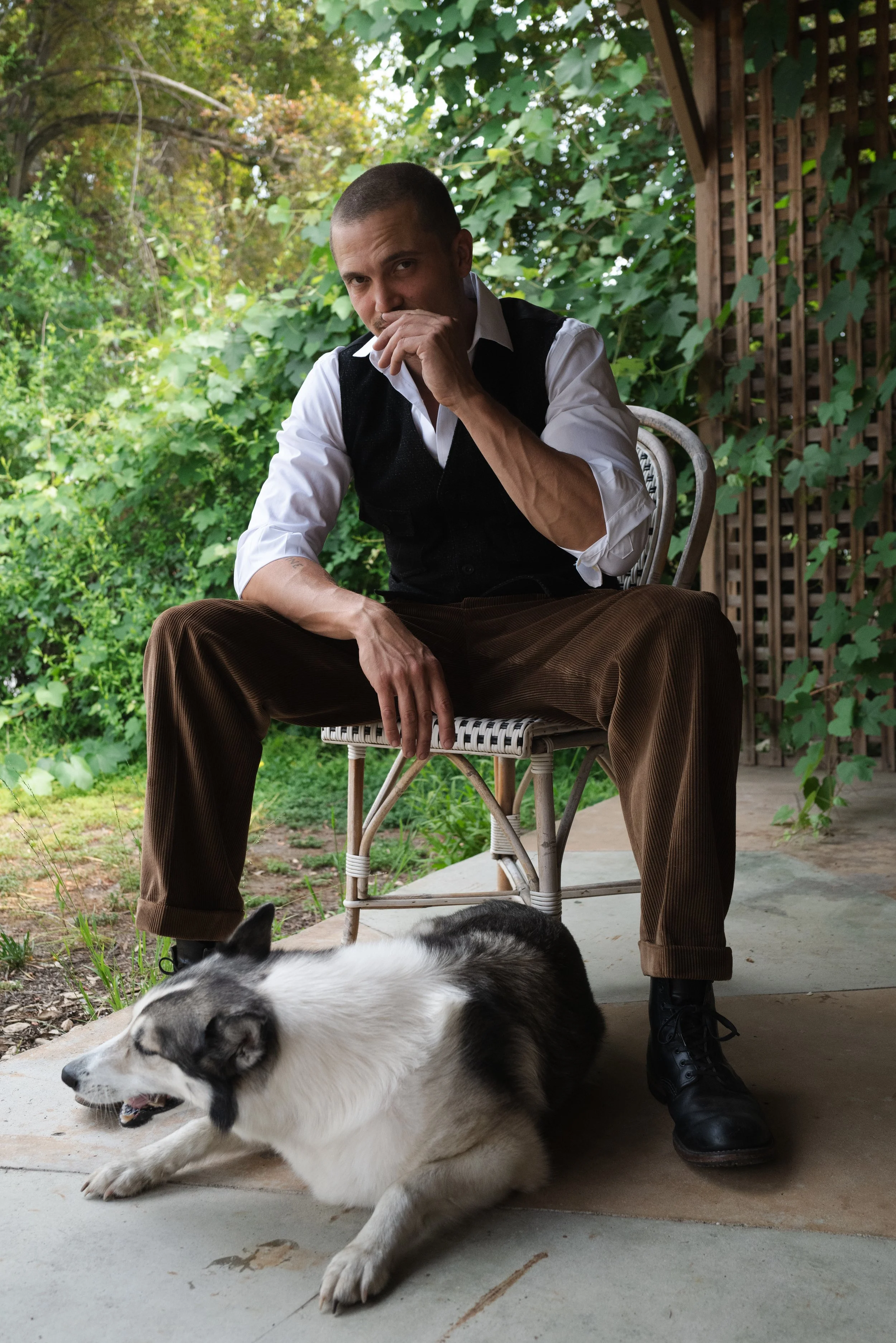 A man sitting outside on a patio, with a husky dog lying on the ground in front of him. The man has a shaved head, wears a white shirt, a black vest, and brown pants, and is touching his face thoughtfully. There are green trees and bushes in the back