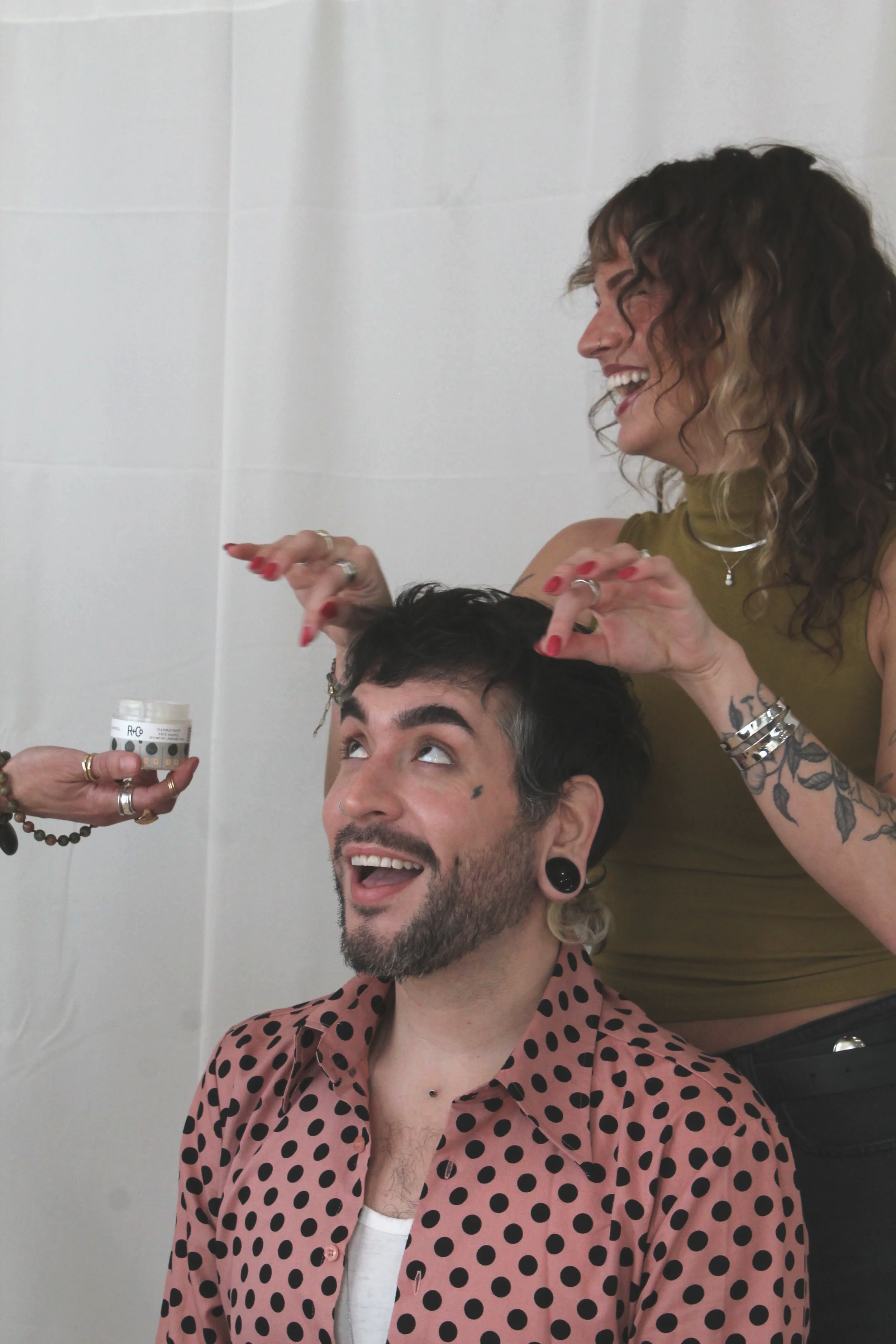 A girl with curly hair in a green shirt applying hair product to the hair of a man in a pink and black polka dot shirt.