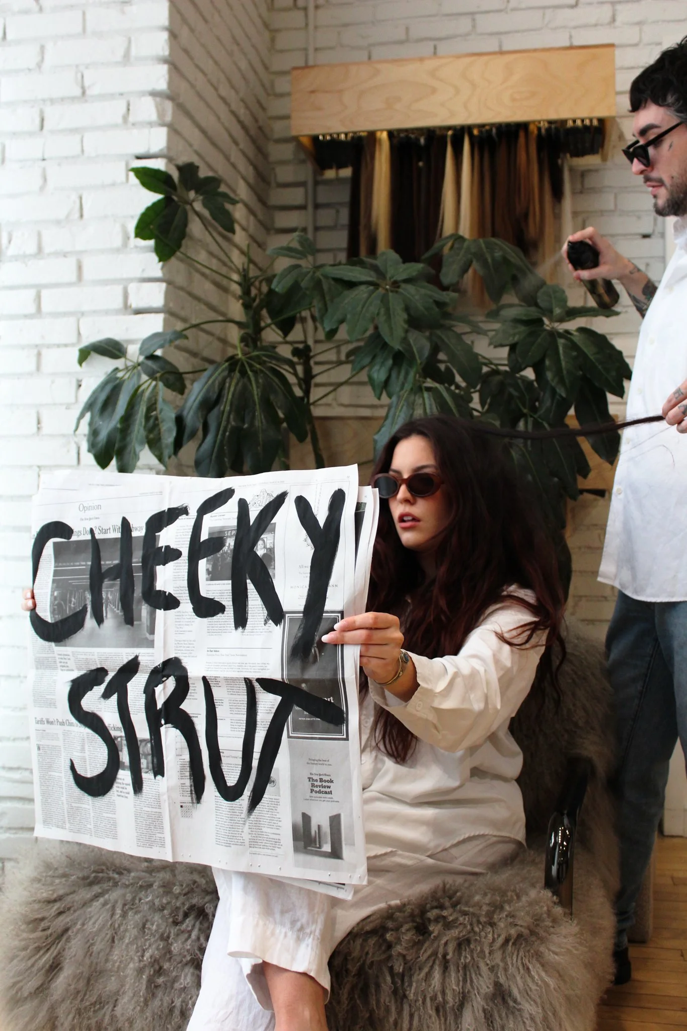 Woman with dark brown hair and sunglasses sitting on a chair reading a newspaper that says "Cheeky Strut."