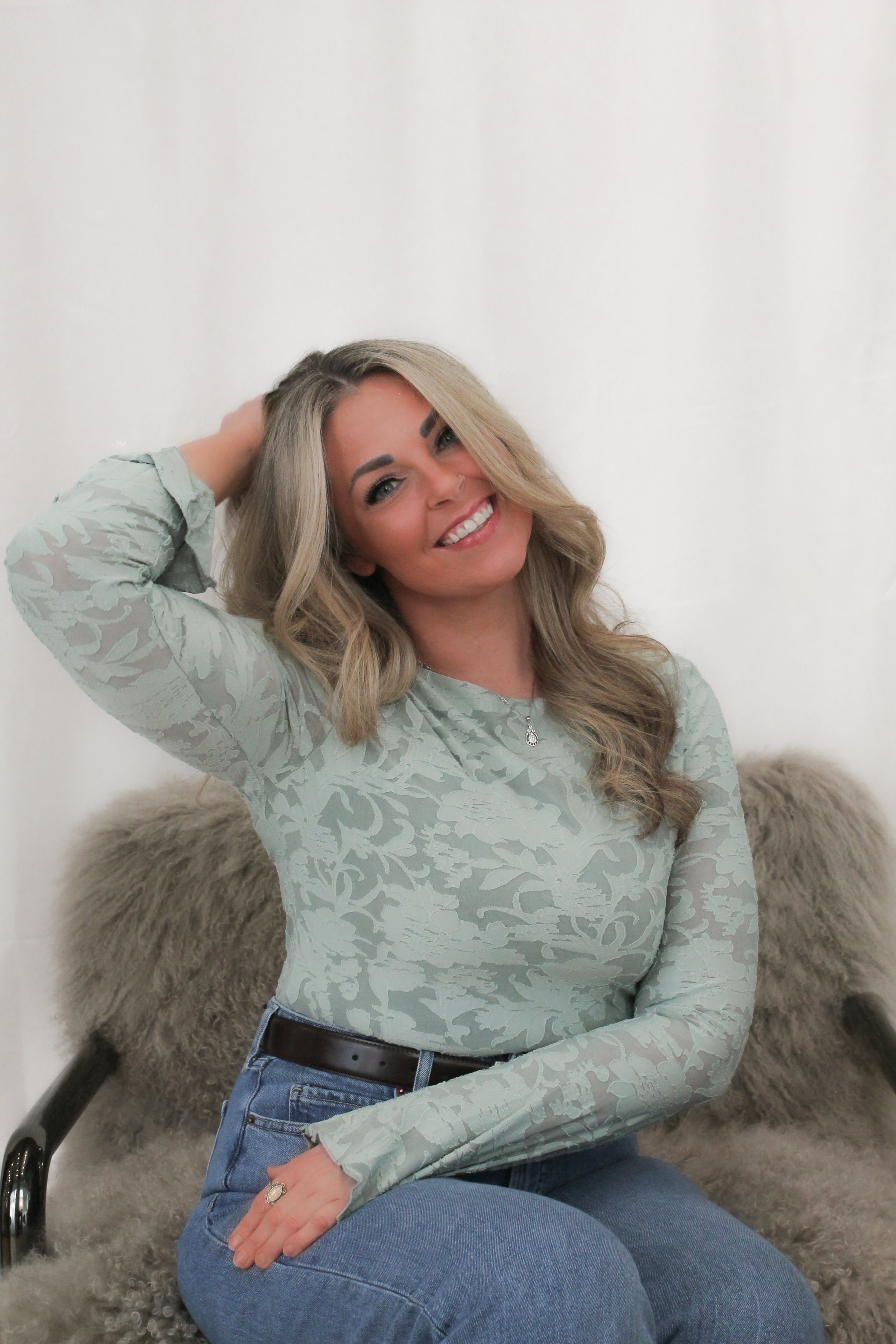 Woman with blonde hair, smiling with a hand in her hair, and sitting on a fur chair.