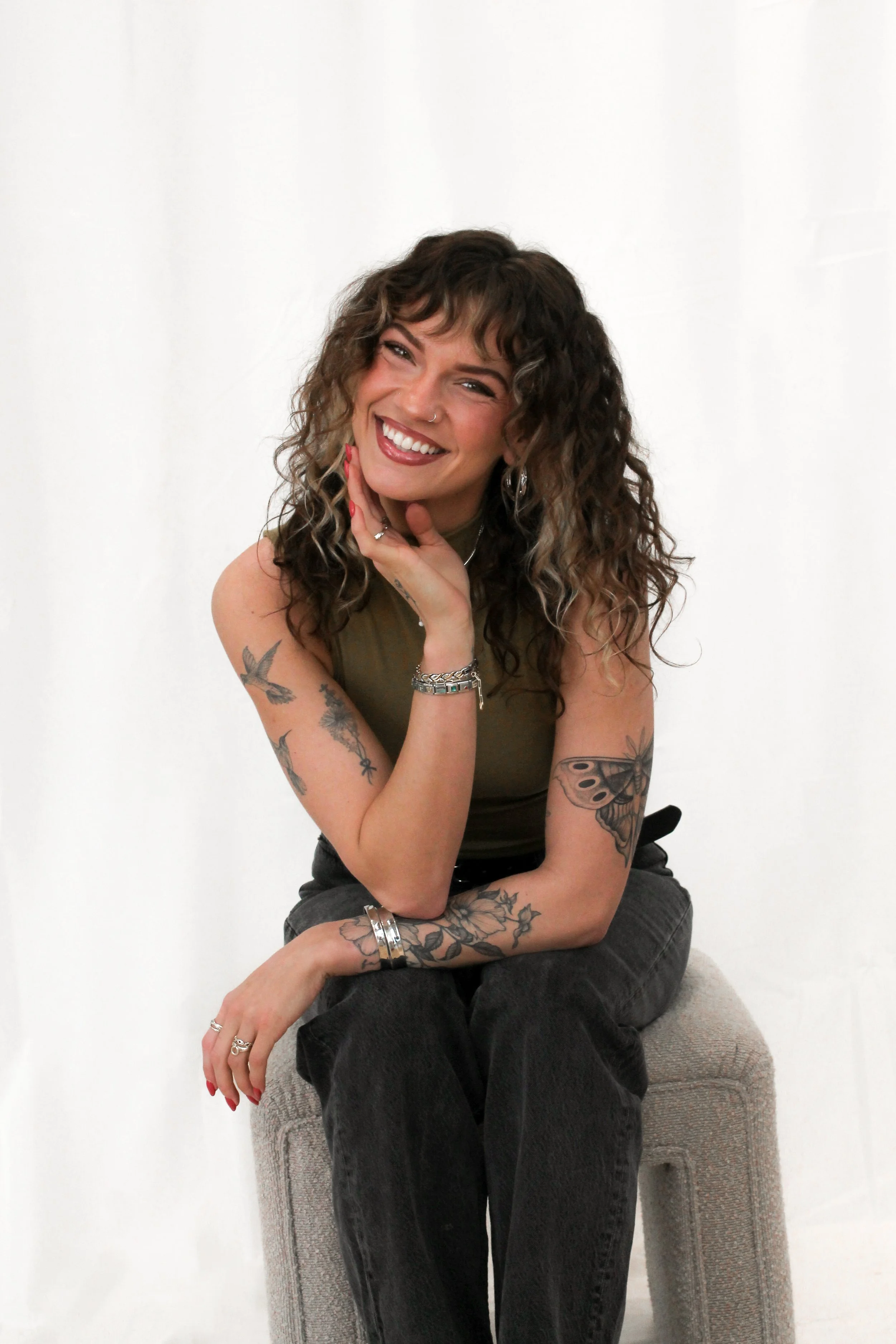 Woman with curly hair sitting on a stool smiling at the camera.