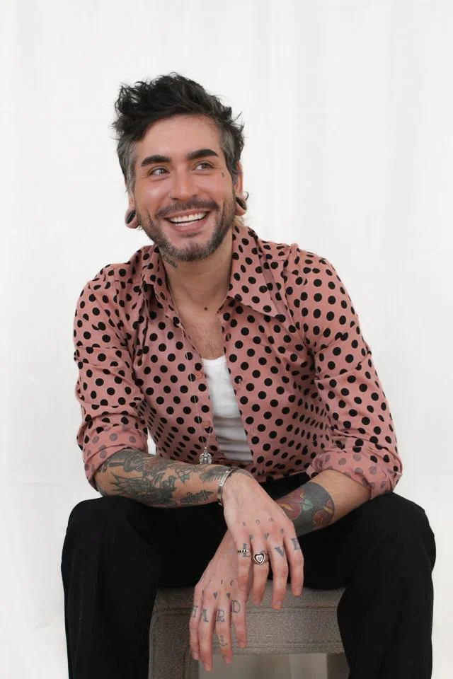 Man sitting in pink and white polka dot shirt and black pants looking to the side.