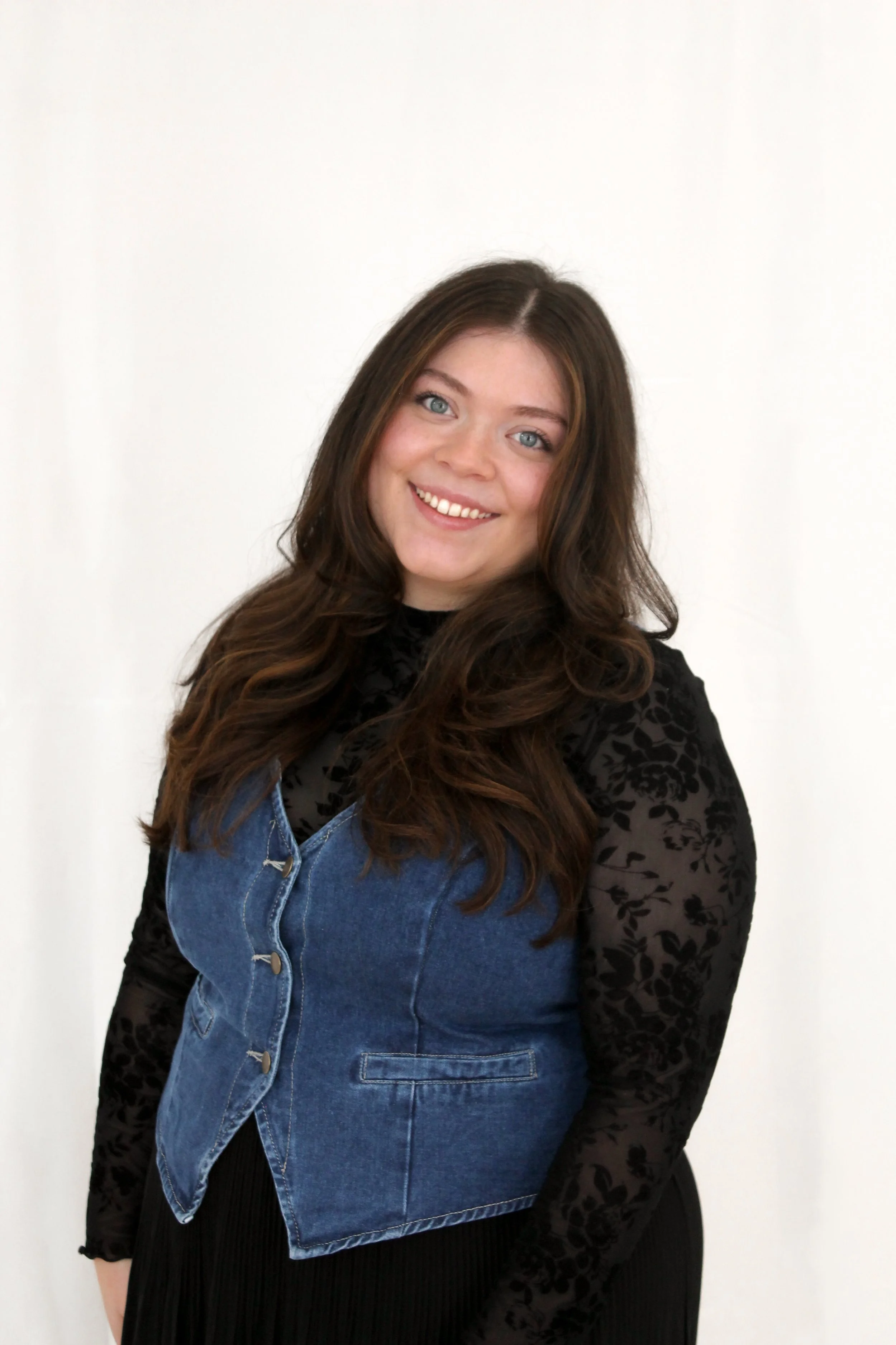 Woman with brown hair in a black long sleeve and denim vest smiling at the camera.