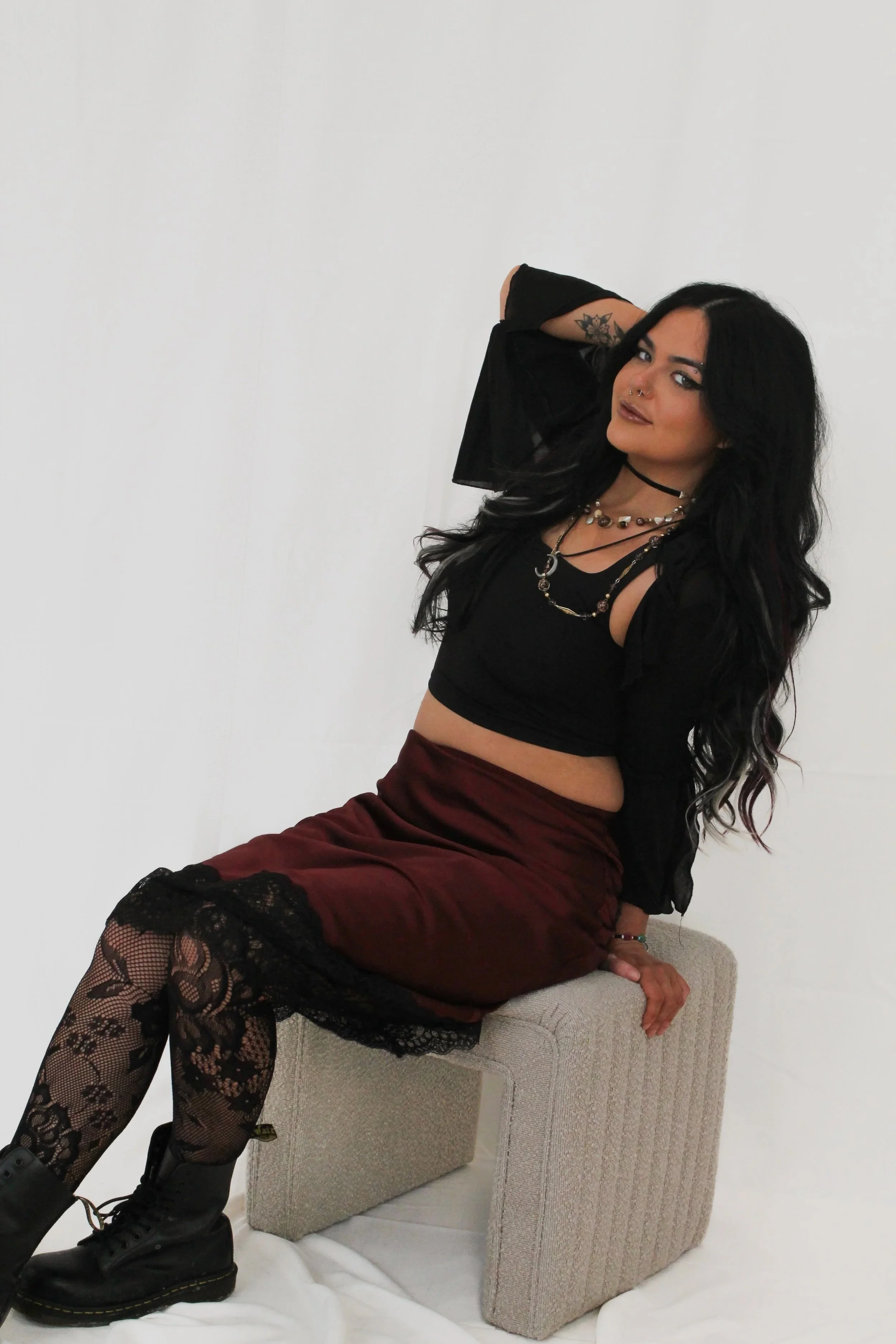 Woman with long black hair and a goth vibe is wearing a black top, burgundy red skirt with lace tights, and combat boots. 