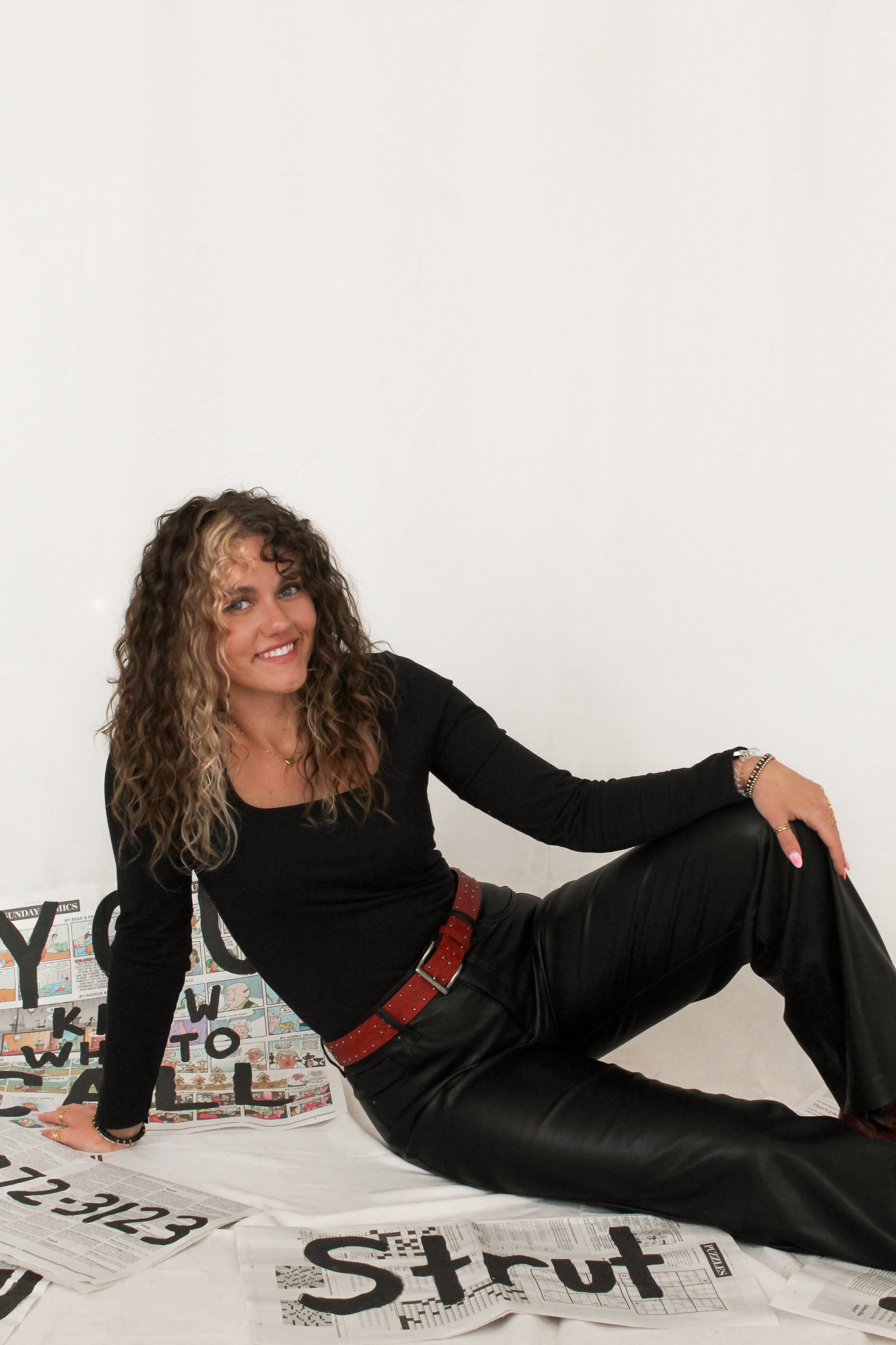 Girl in all black with brown curly hair sitting on the ground smiling