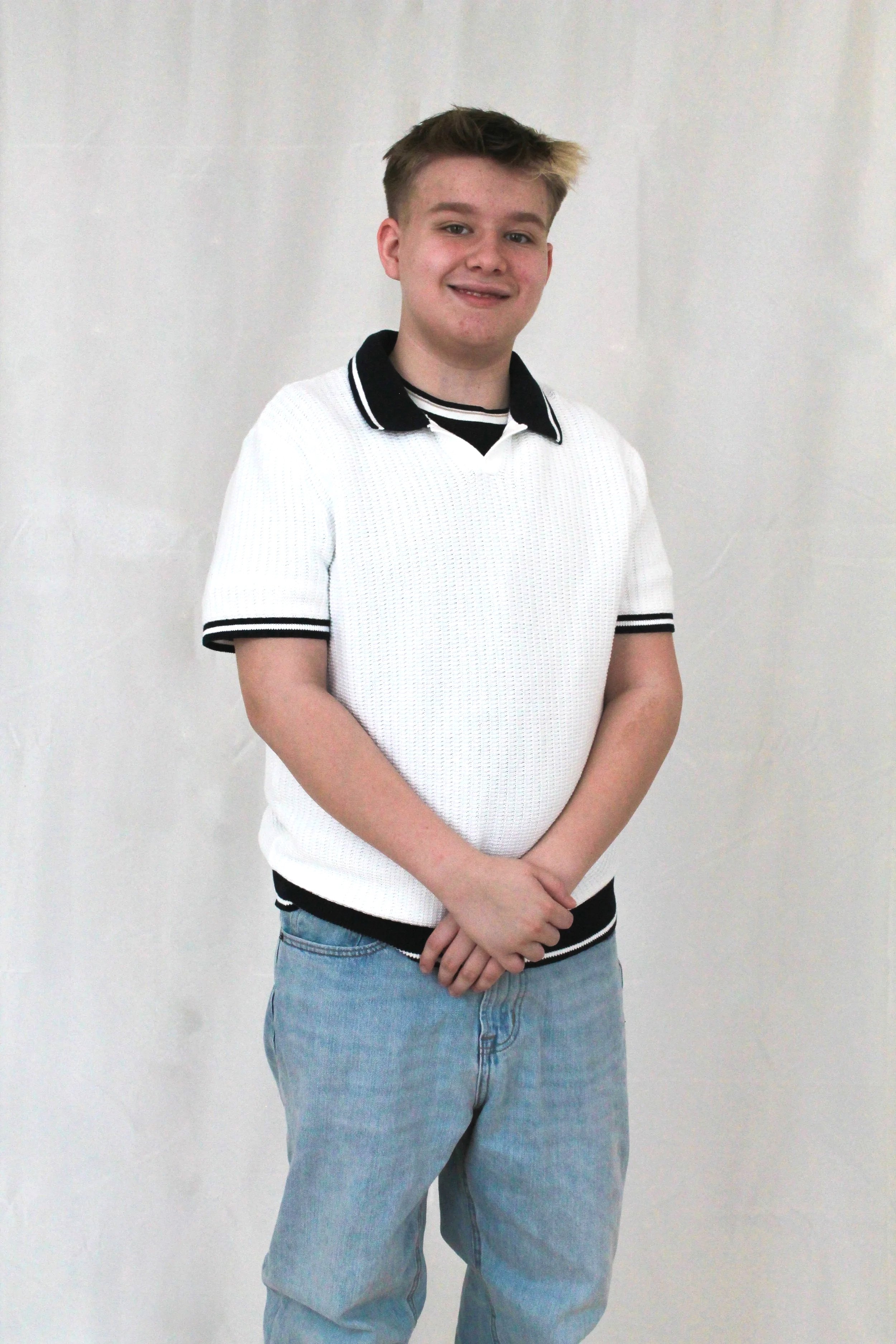 Boy with short blonde hair in a white shirt standing and smiling at the camera.