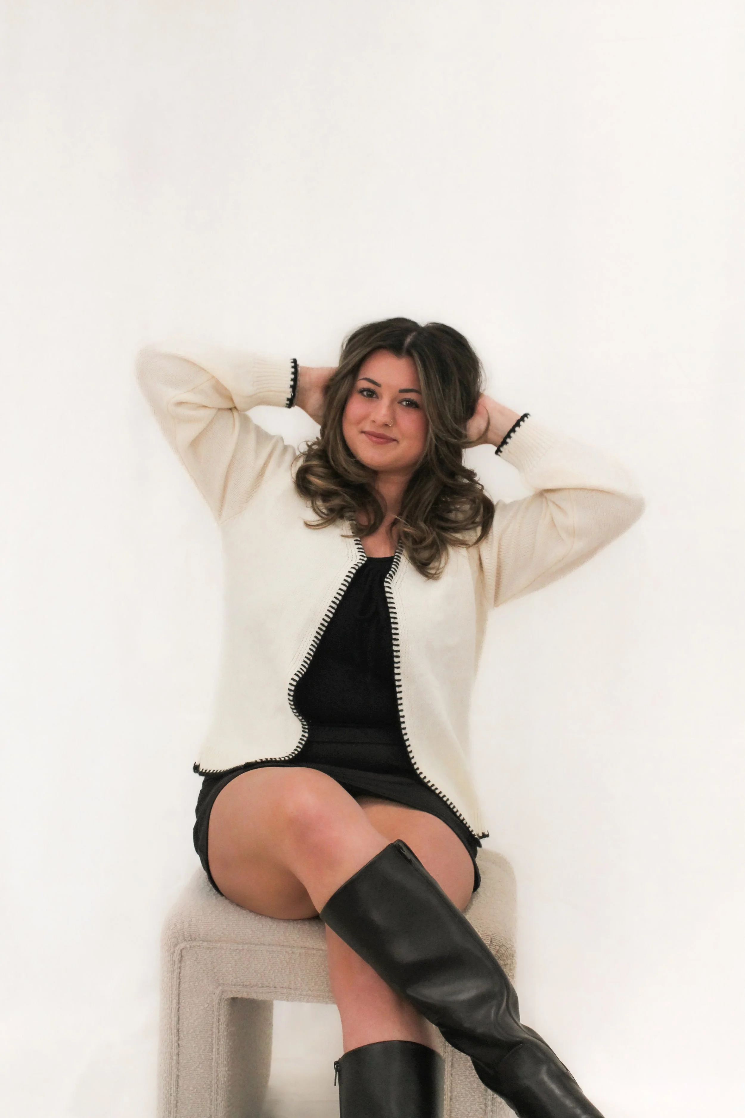 Woman with dark brown hair wearing a white sweater and black dress, while sitting cross legged and her hands in her hair. 