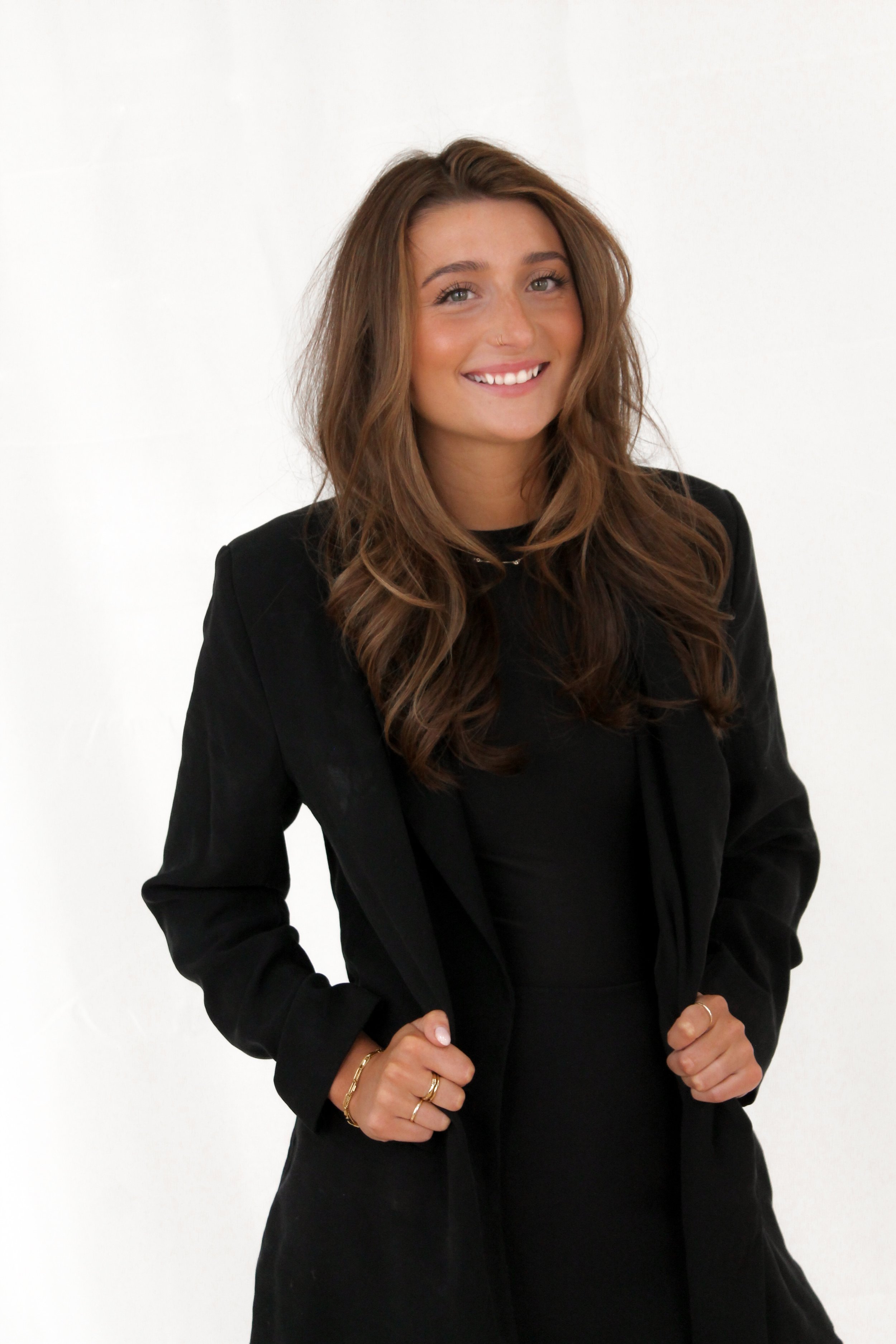 Woman with dark brown highlighted hair, wearing a black dress and black blazer.