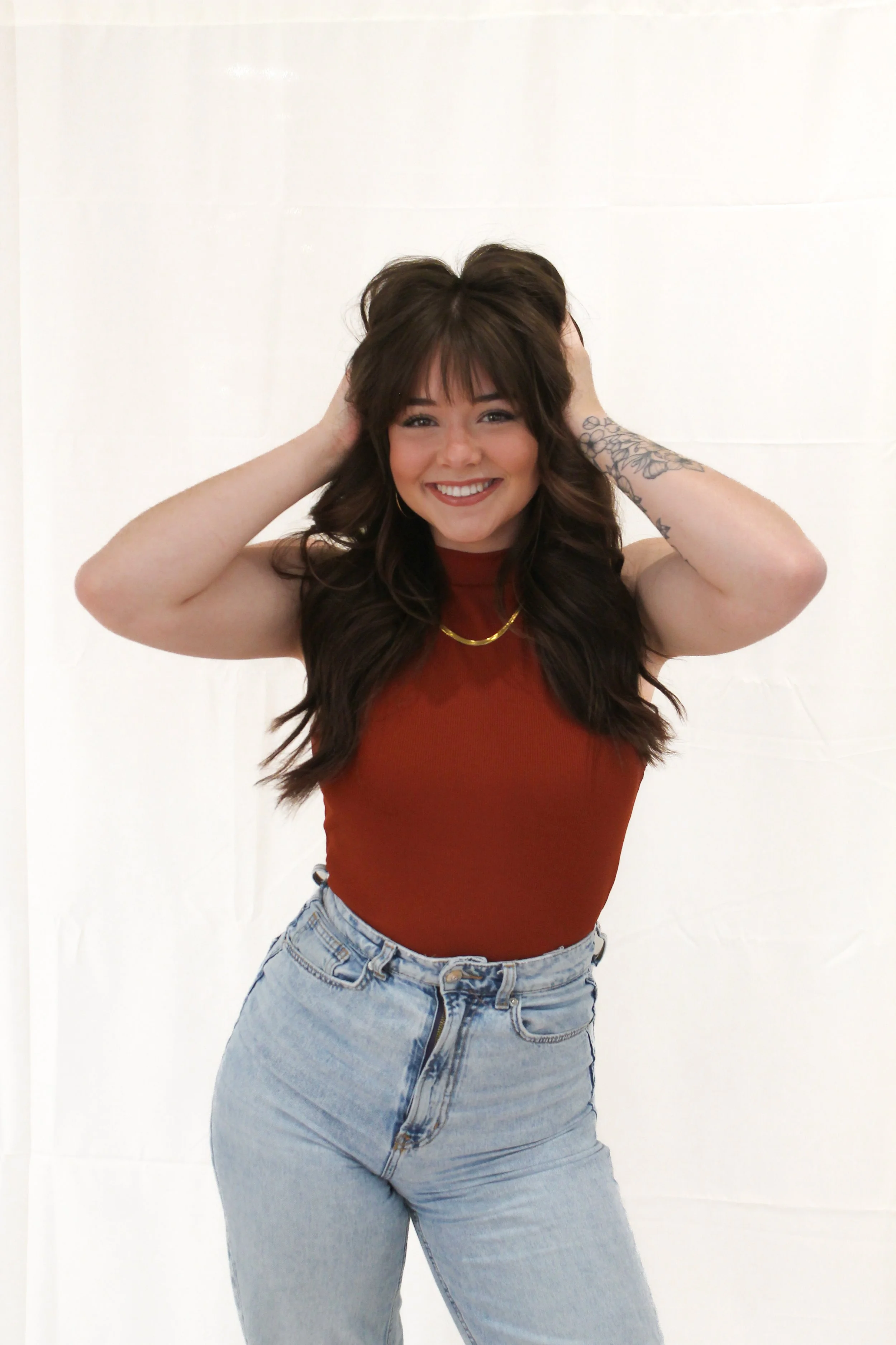 Woman smiling with curled dark brown hair, wearing a red top with her hands in her hair. 