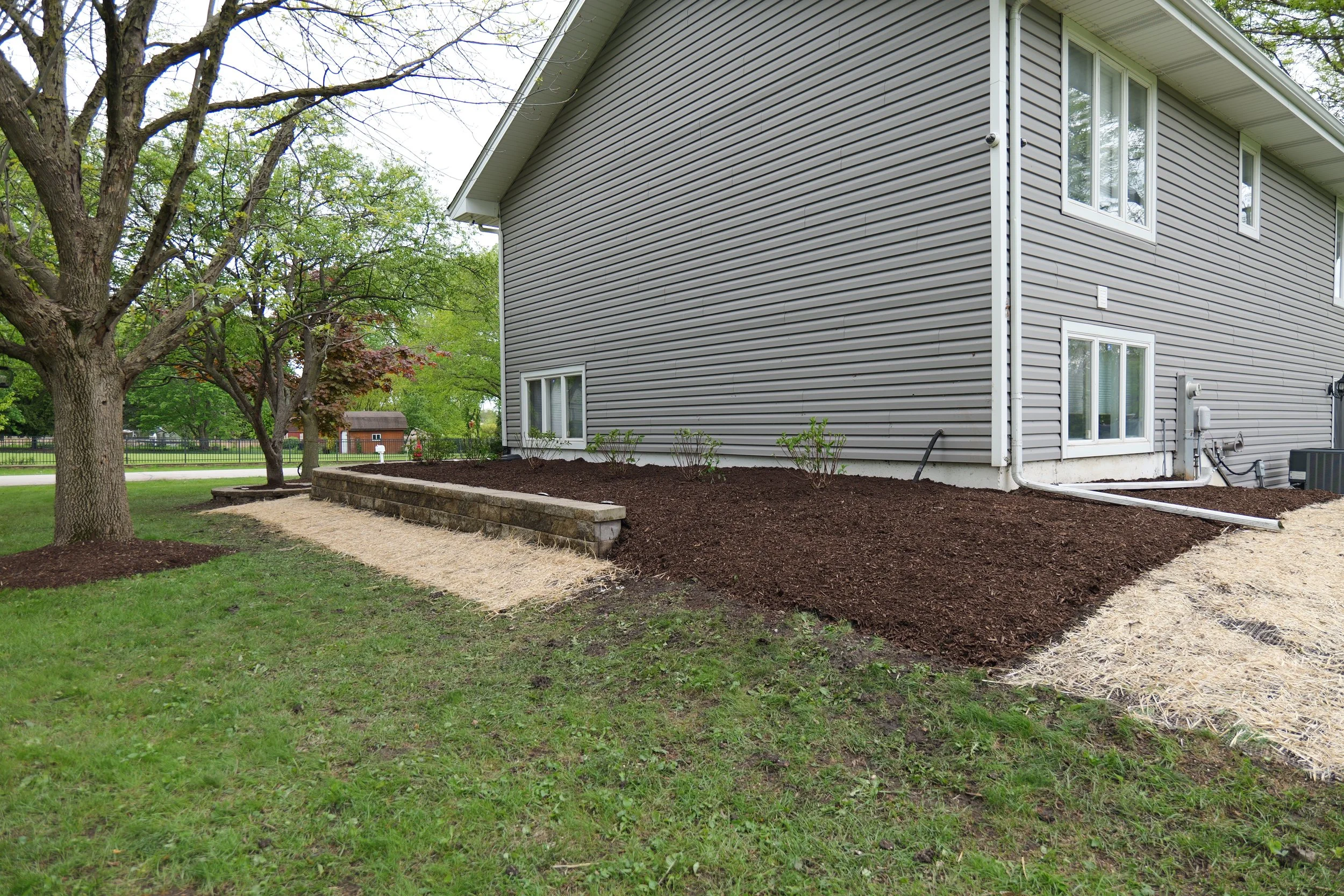 A house with gray vinyl siding, several windows, and a landscaped yard with a tree, mulch, and a stone border.