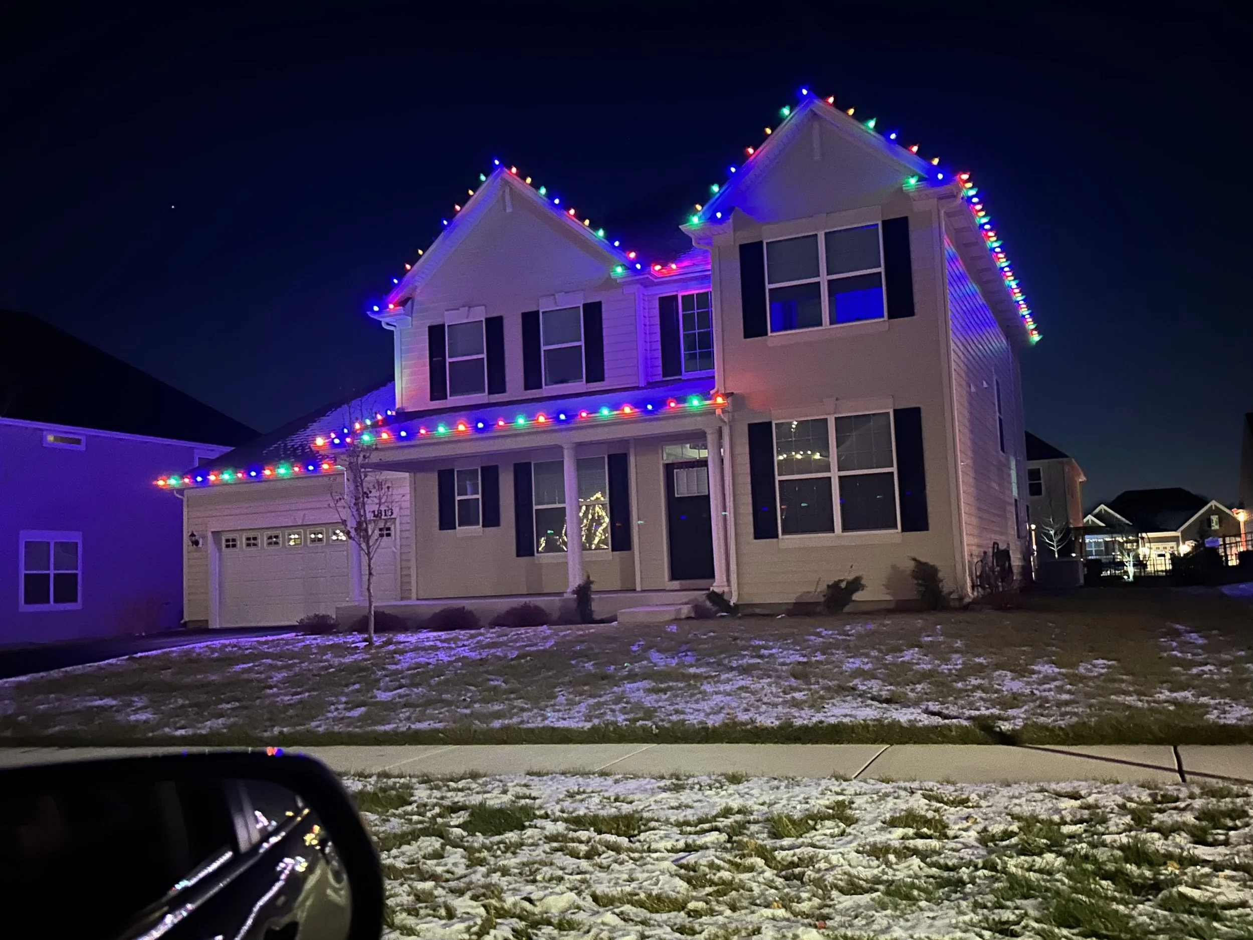 A two-story house decorated with colorful Christmas lights on the roof and along the edges, with snow-covered grass in the front yard at night.