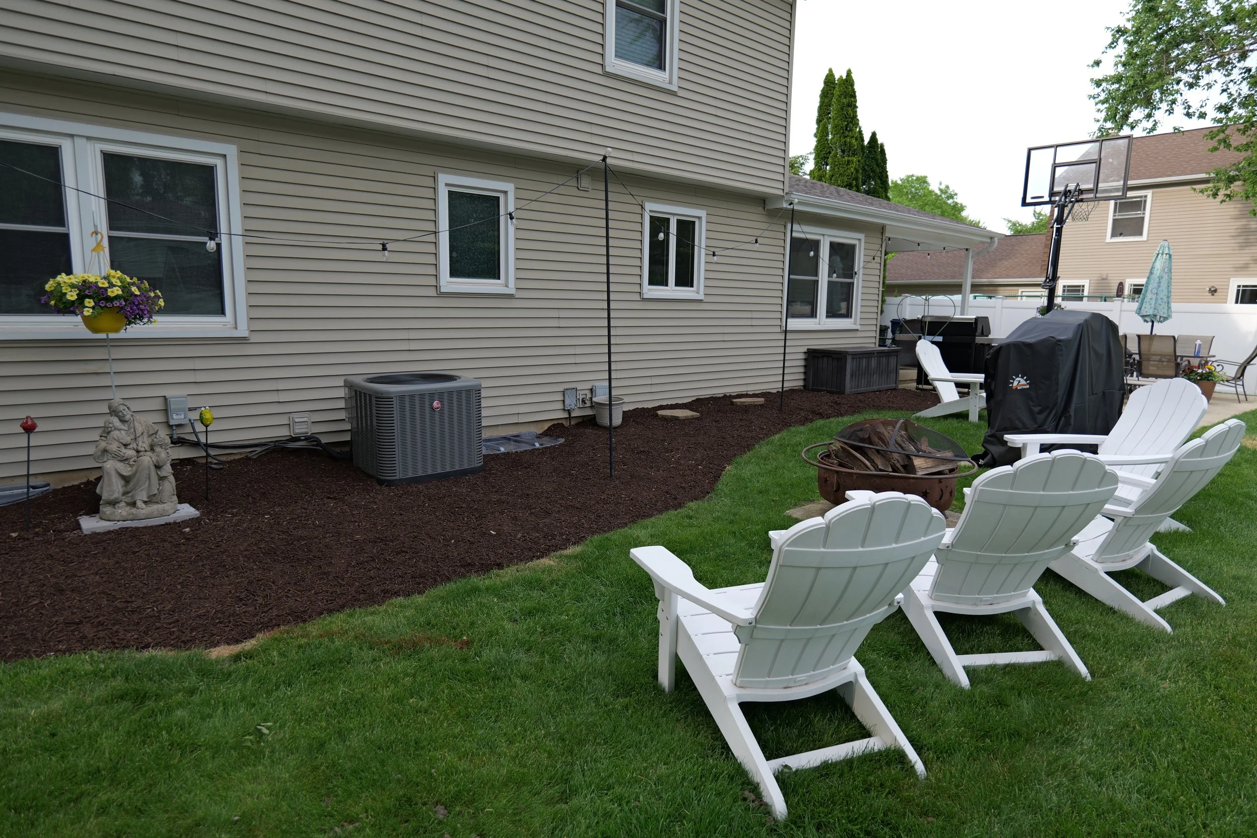Backyard with patio chairs, a firepit, a covered grill, a garden statue, hanging flower basket, string lights, and a basketball hoop.