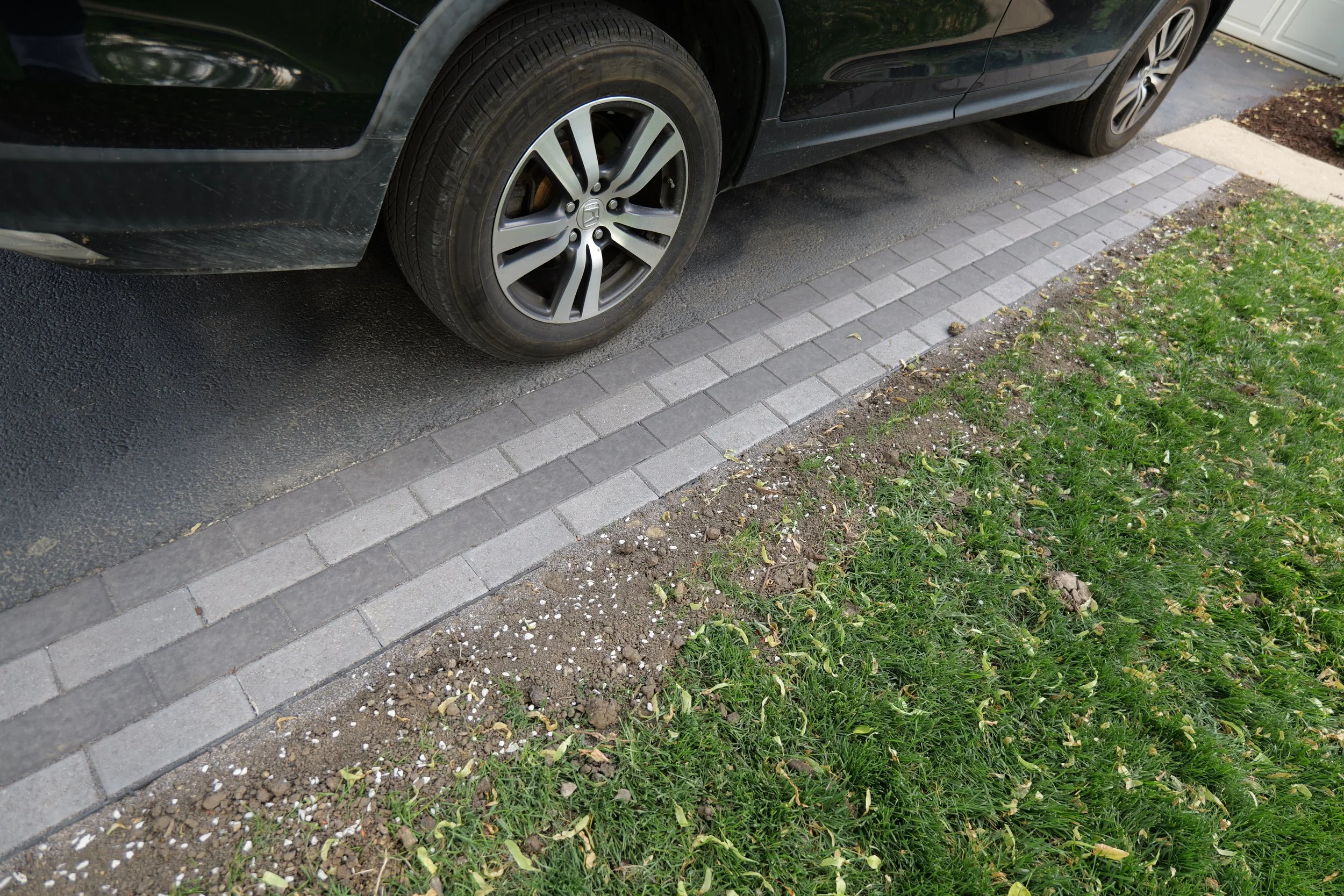 A black car parked on a driveway with brick paving, next to a grassy lawn and garden bed with some yellow flowers and green plants.