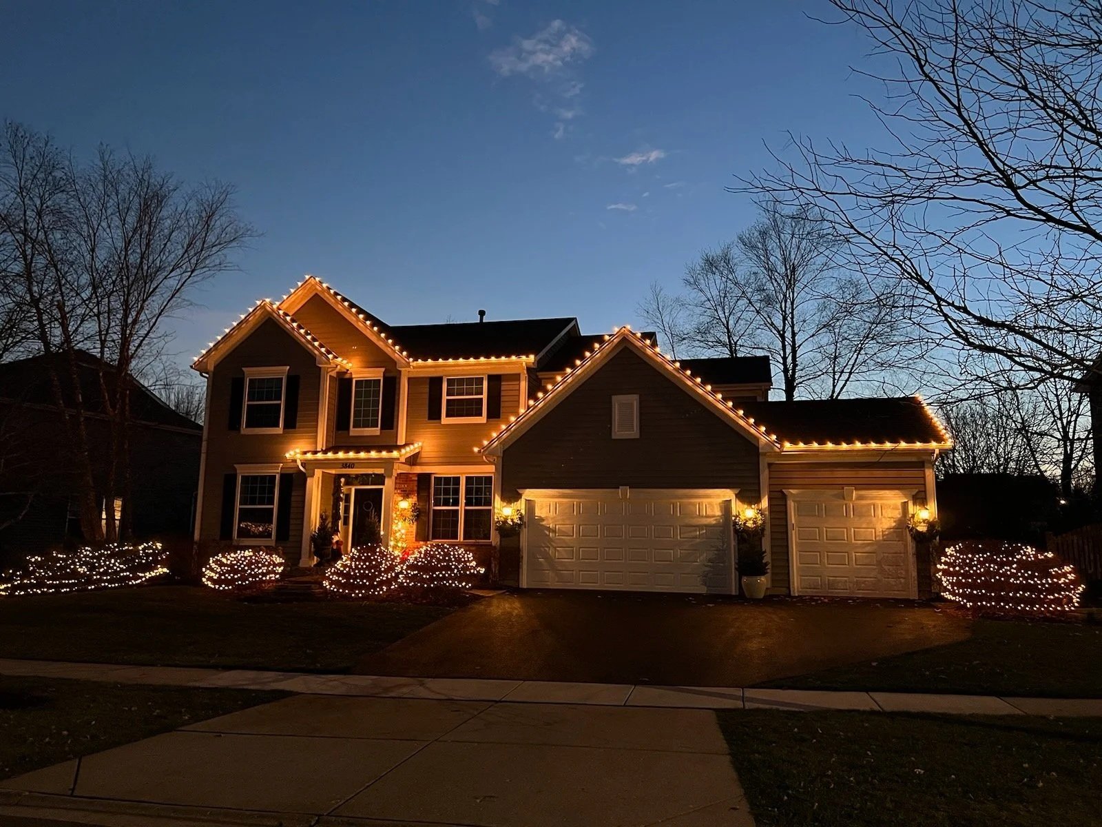 A house decorated with Christmas lights in the evening, with lights outlining the roof and bushes in the yard.