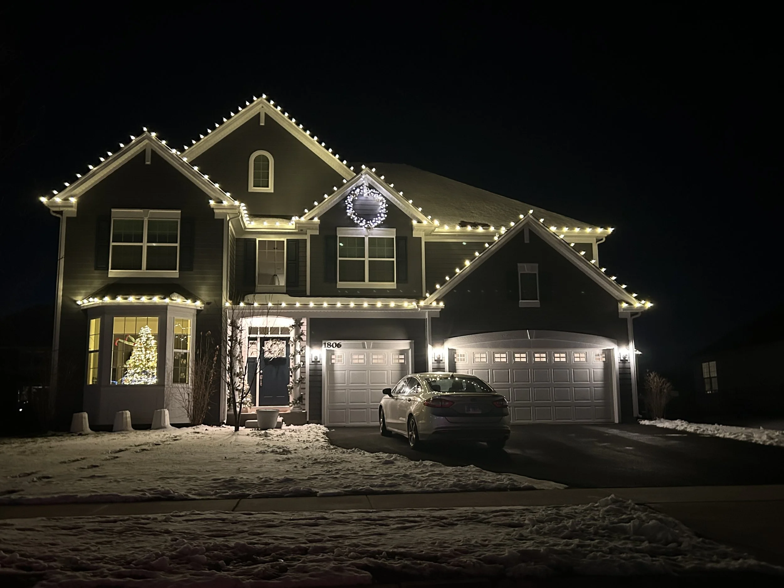 A house decorated with Christmas lights at night, featuring a wreath on the front of the house, a Christmas tree visible through a window, and a car parked in the driveway. Snow covers the ground.