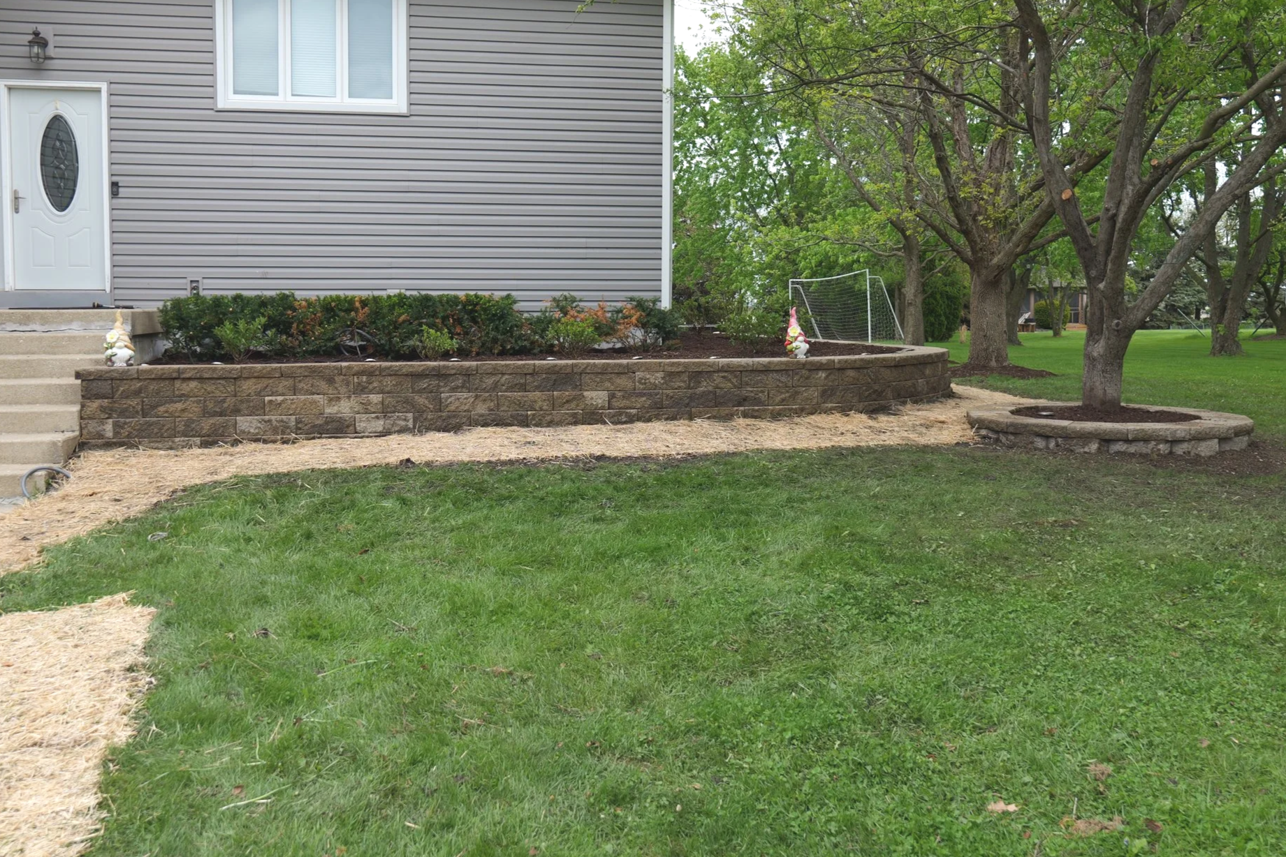 A backyard garden with a raised stone flower bed, decorative gnomes, a large tree with a circular stone border, and a grassy lawn.