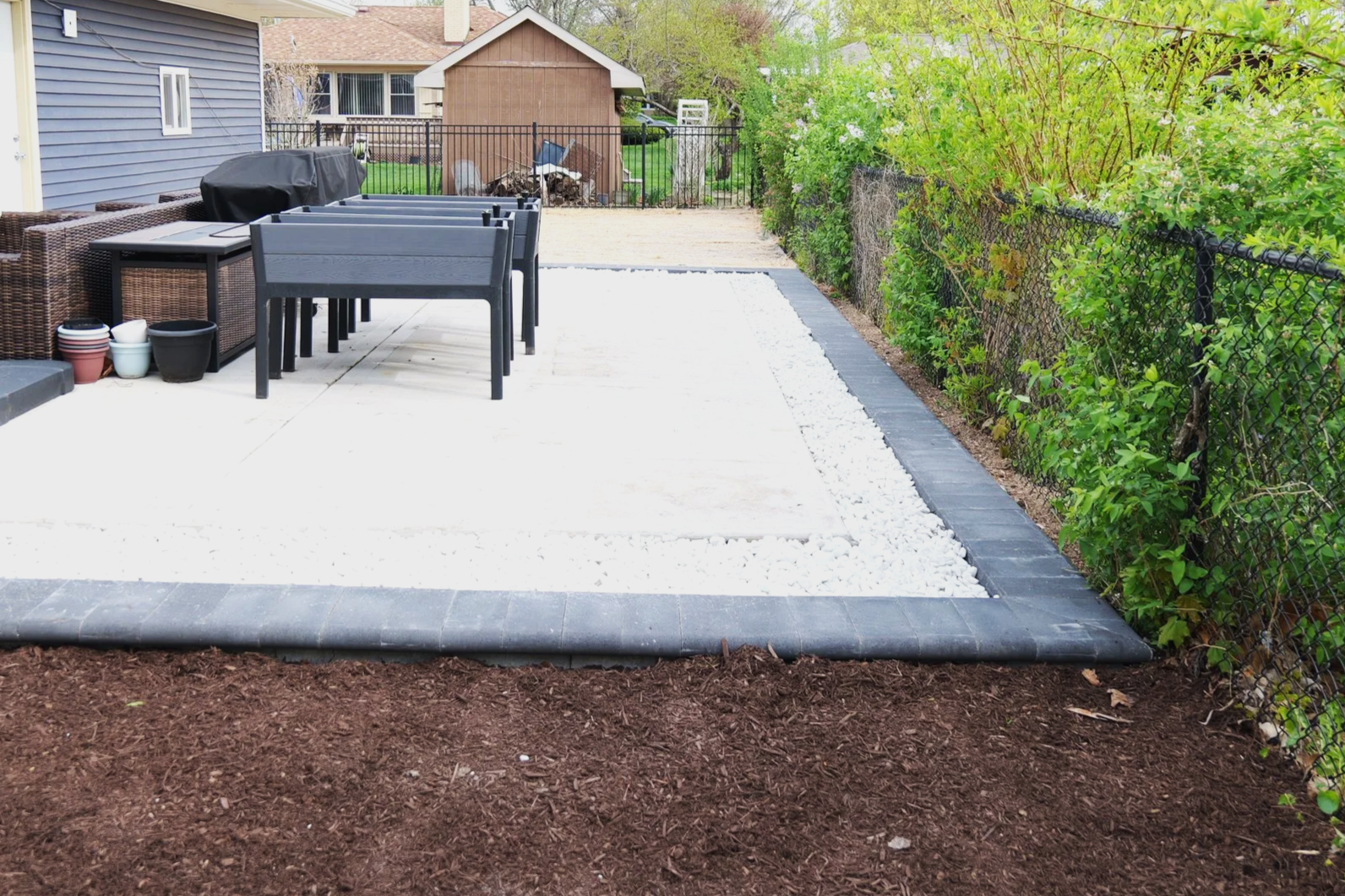 Backyard patio with a stone border, tables, chairs, plants, and a fenced yard with a shed and trees.