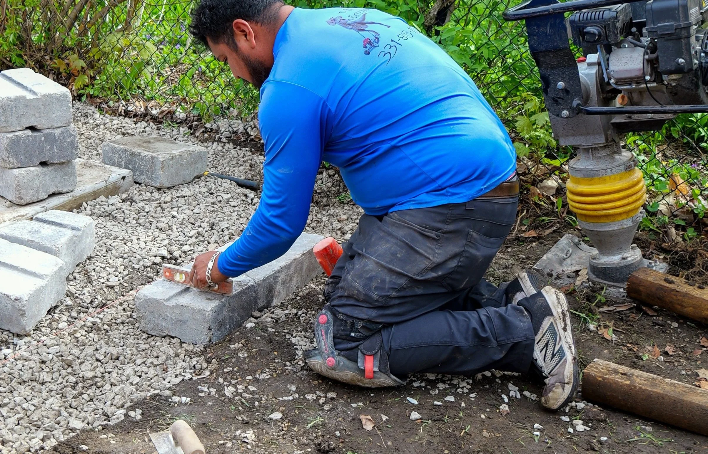 A man kneels on the ground and uses a level to work on laying brick pavers in a garden or yard. He is dressed in a bright blue hoodie, black pants, and sneakers. There is a radio on the right side of the image and a chain-link fence with greenery behind it.