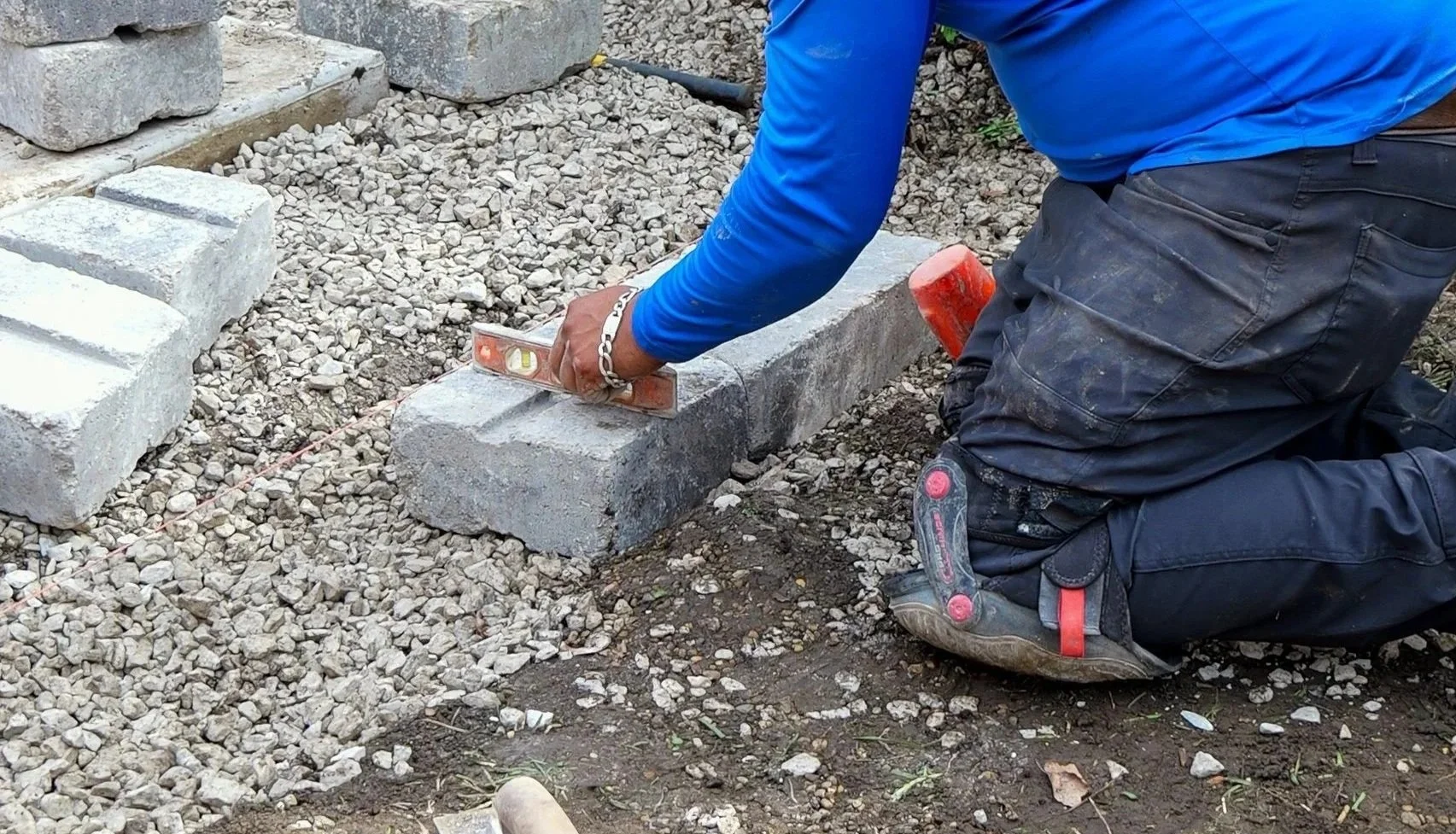A worker kneeling on the ground lays bricks, holding a level in one hand and a hammer in the other, working on a brick pathway or wall.