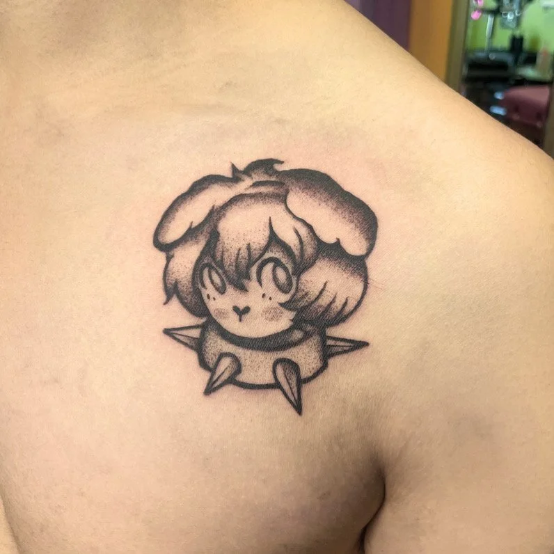 A black and gray tattoo of a cute, cartoon-style puppy with floppy ears and a collar with spikes on a person's shoulder.